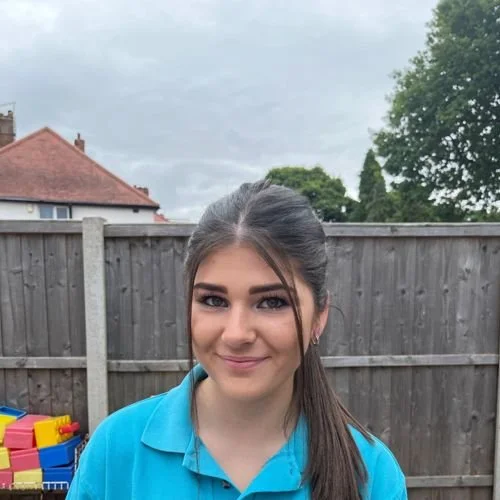 Young woman with dark hair in a ponytail smiling outdoors in front of a wooden fence and cloudy sky.