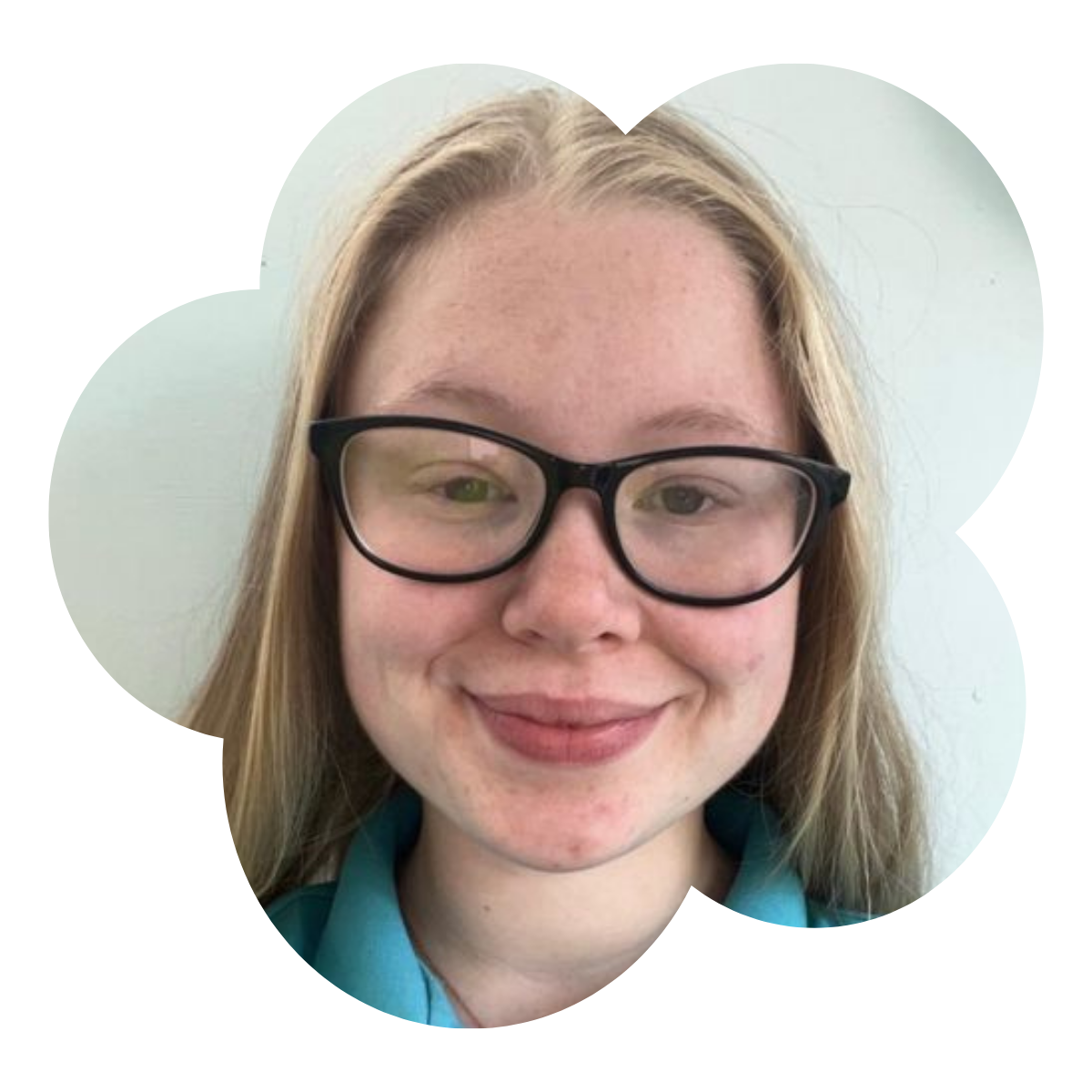 A young woman with long blonde hair and glasses taking a selfie in front of a light green wall.