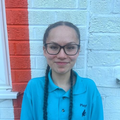 A young girl with glasses and braided hair standing in front of a painted brick wall, wearing a blue uniform shirt.