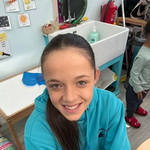 A warm and friendly member of nursery staff smiling in a smart uniform in a pre-school classroom in a childcare nursery in Solihull.