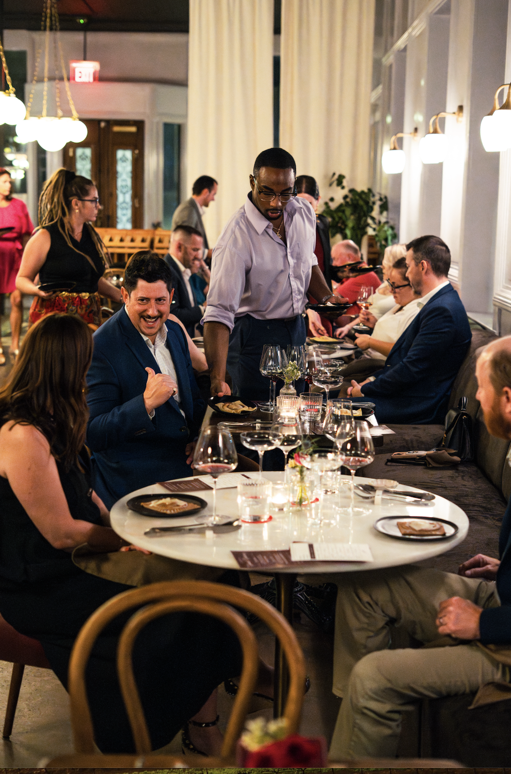 Groups of people dining at a restaurant with servers serving food, elegant table settings, wine glasses, and a cozy, well-lit atmosphere.