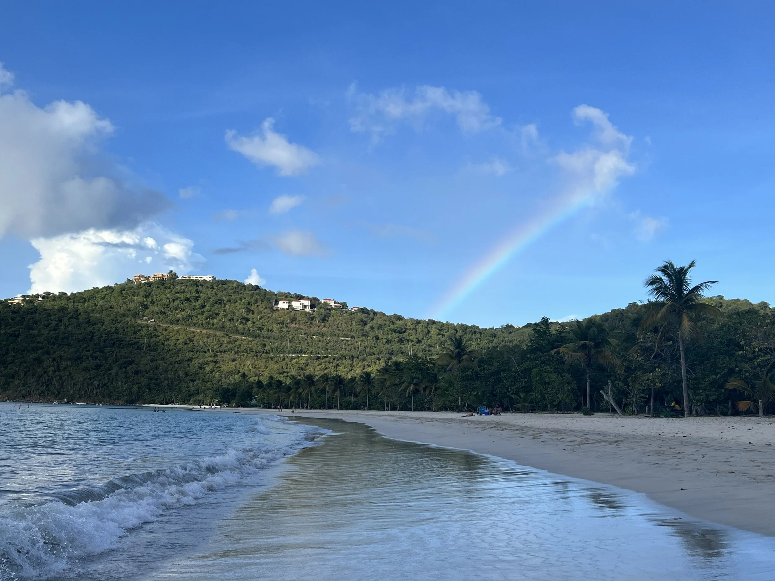 Calm beach with gentle waves, sandy shoreline, and tropical palm trees, hillside with houses, blue sky with scattered clouds, and a faint rainbow.