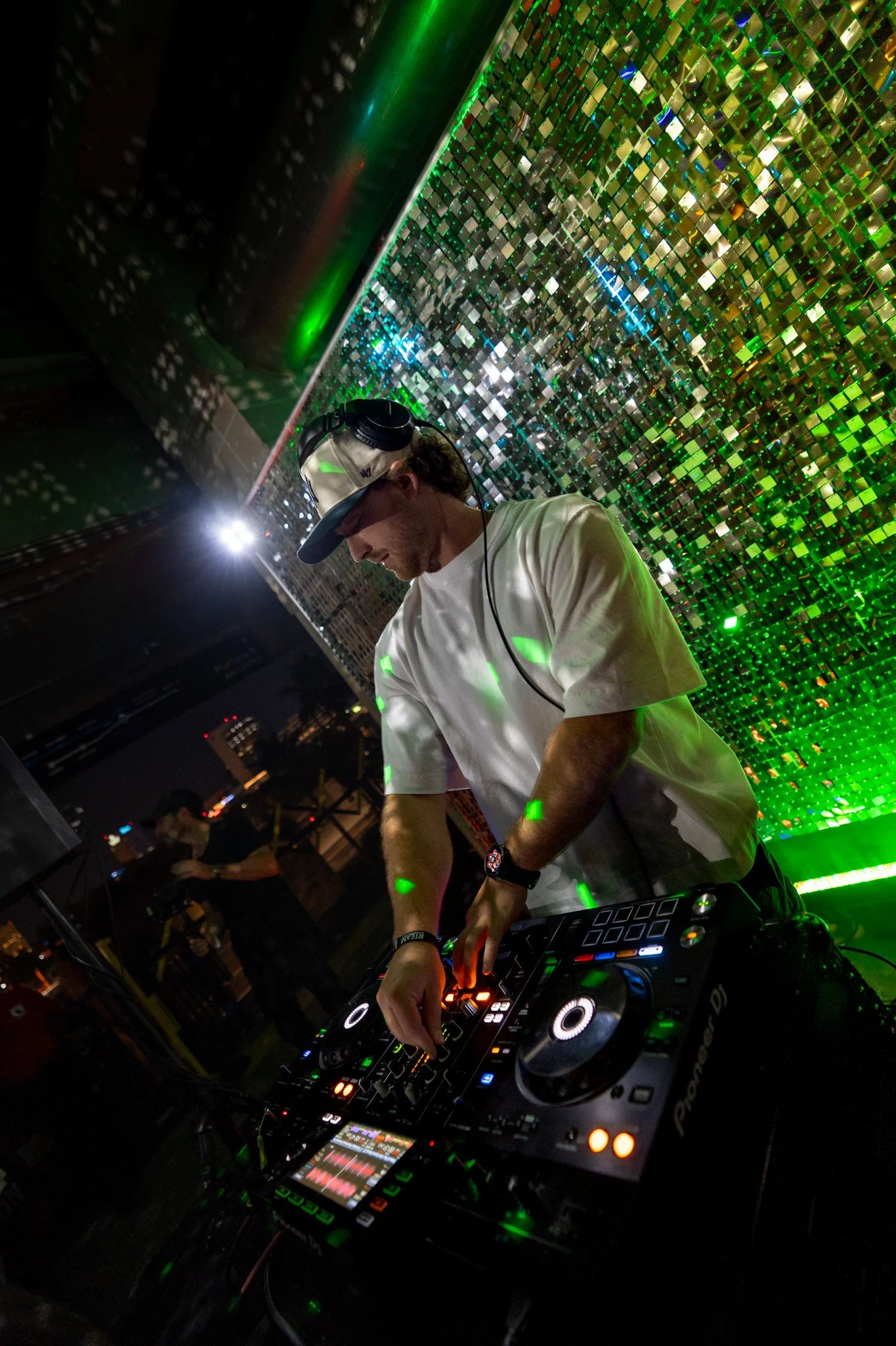 DJ wearing a cap and headphones performing at a nightclub with a reflective, multicolored mosaic backdrop.