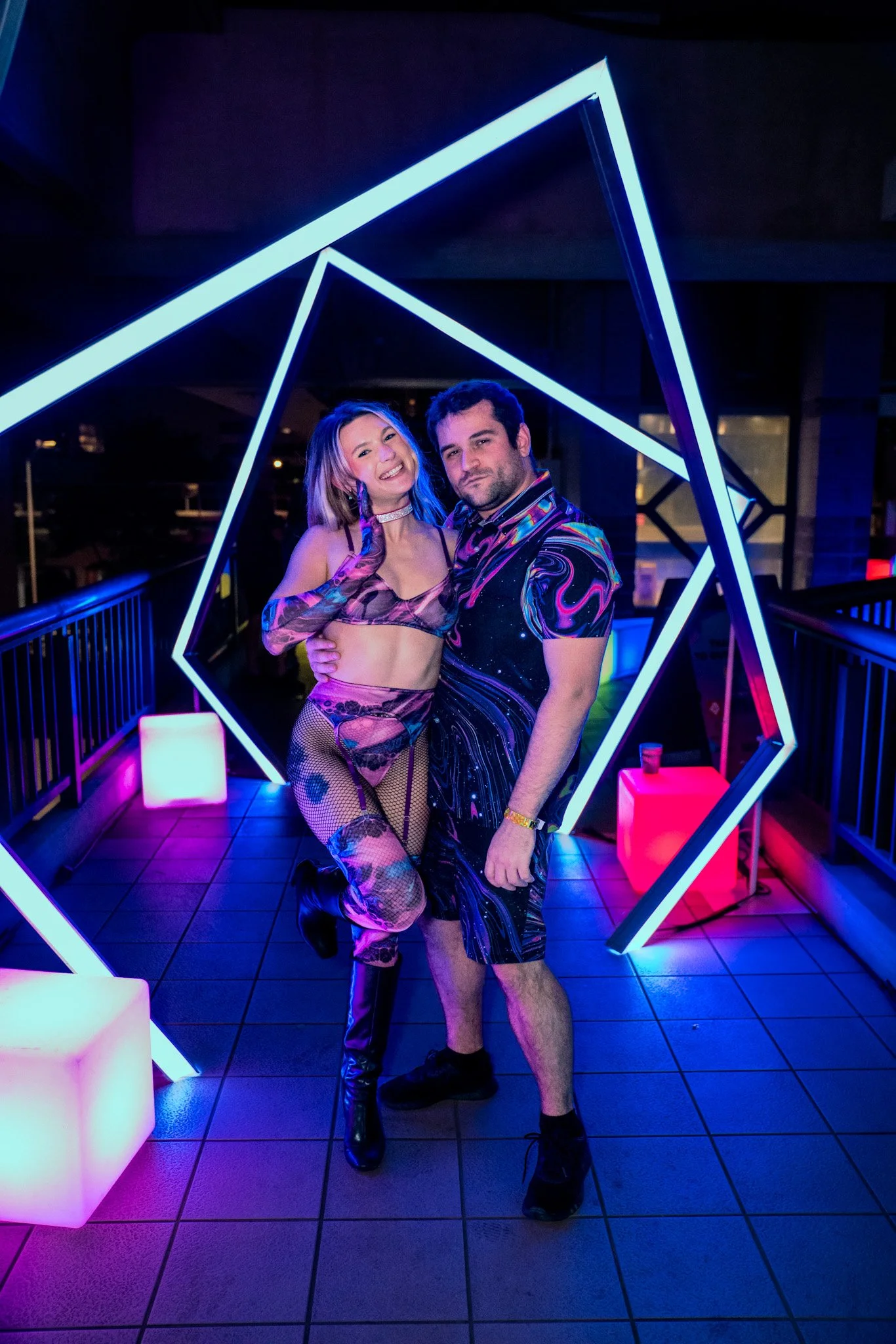 A woman and a man posing together in a neon-lit setting at night, with geometric light structures around them. The woman is wearing a colorful, patterned outfit with fishnet stockings and thigh-high boots, while the man is in a matching swirling patt