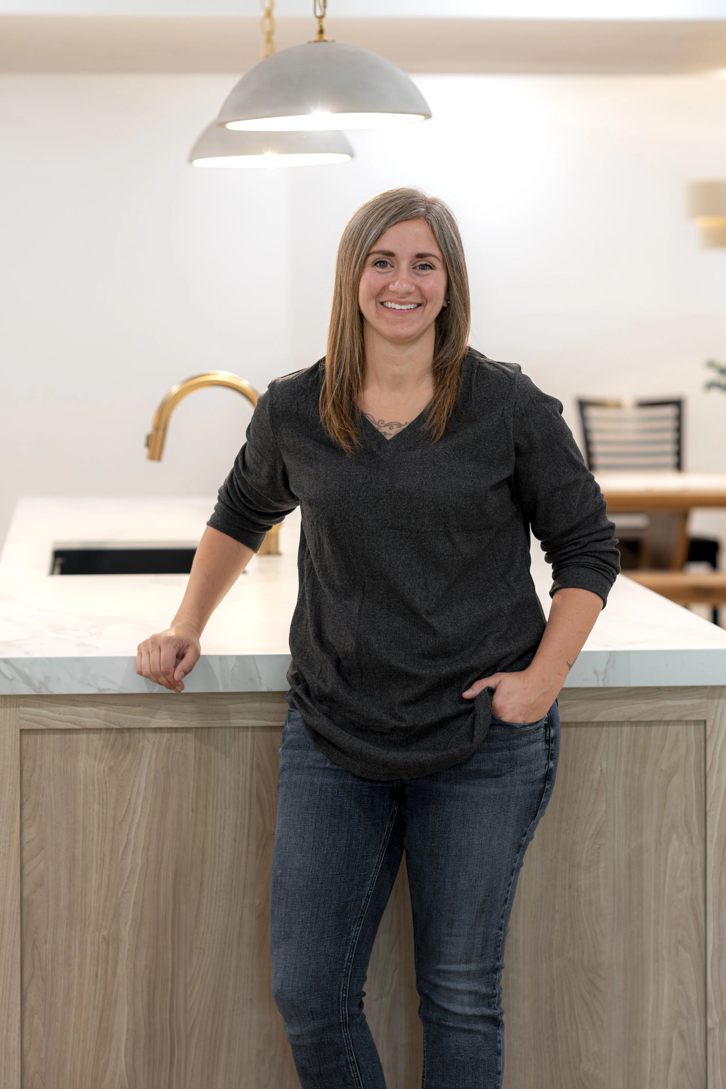 A woman with shoulder-length brown hair, wearing a black long-sleeve top and jeans, standing in a modern kitchen with a marble countertop and gold faucet, smiling at the camera.