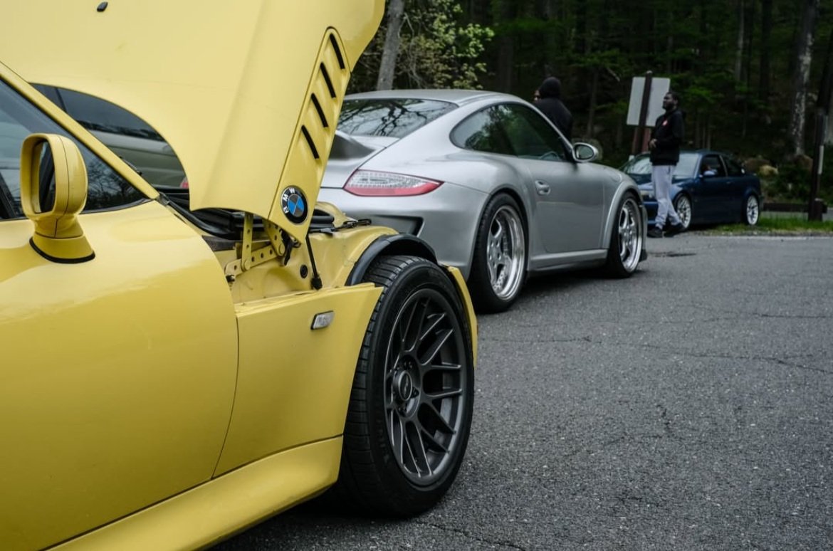Yellow z3 bmw with hood open and a silver porche in the foreground.