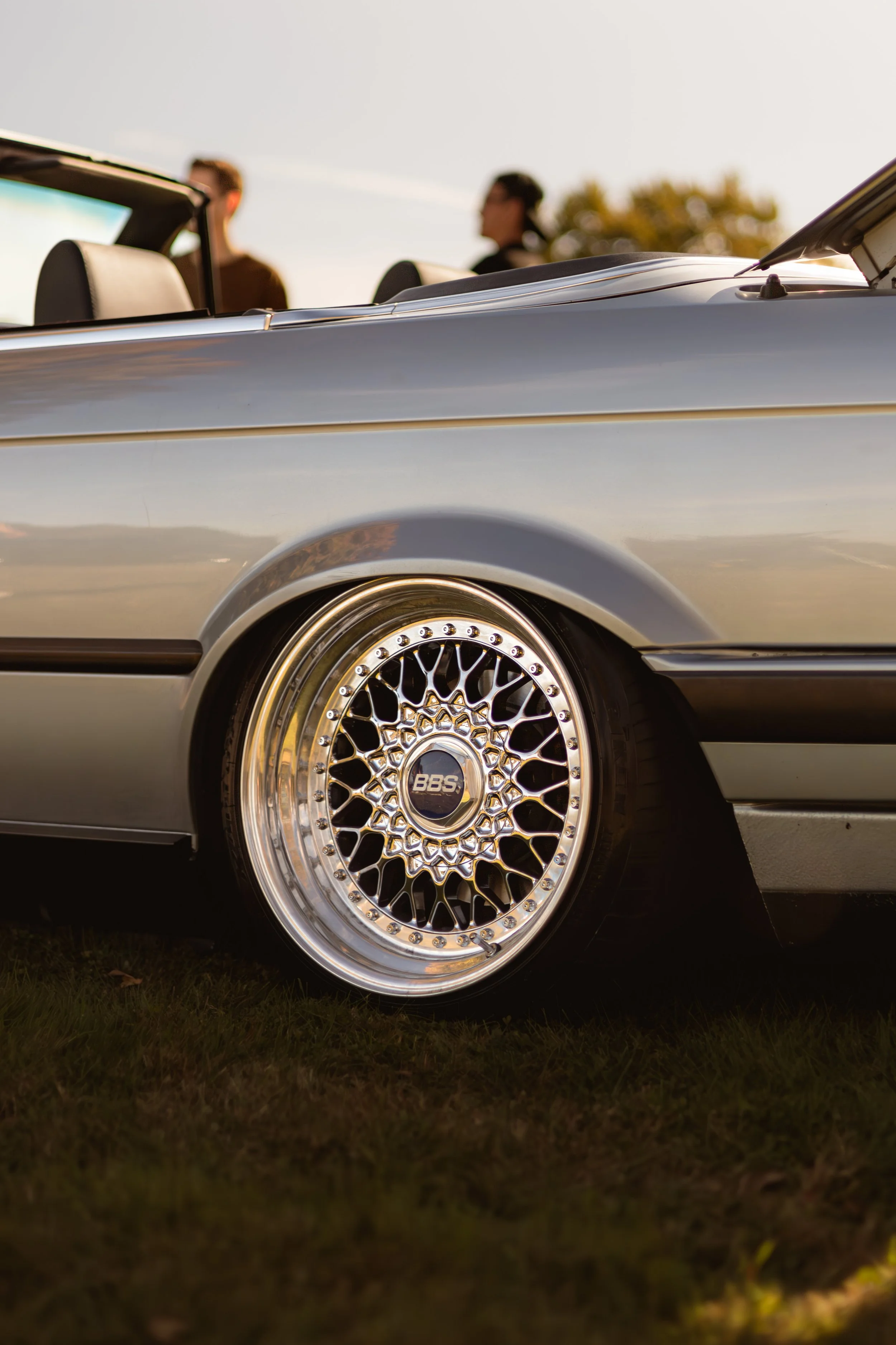 Close-up of a silver convertible car with BBS alloy wheels, parked on grass, with two people standing in the background during sunset.