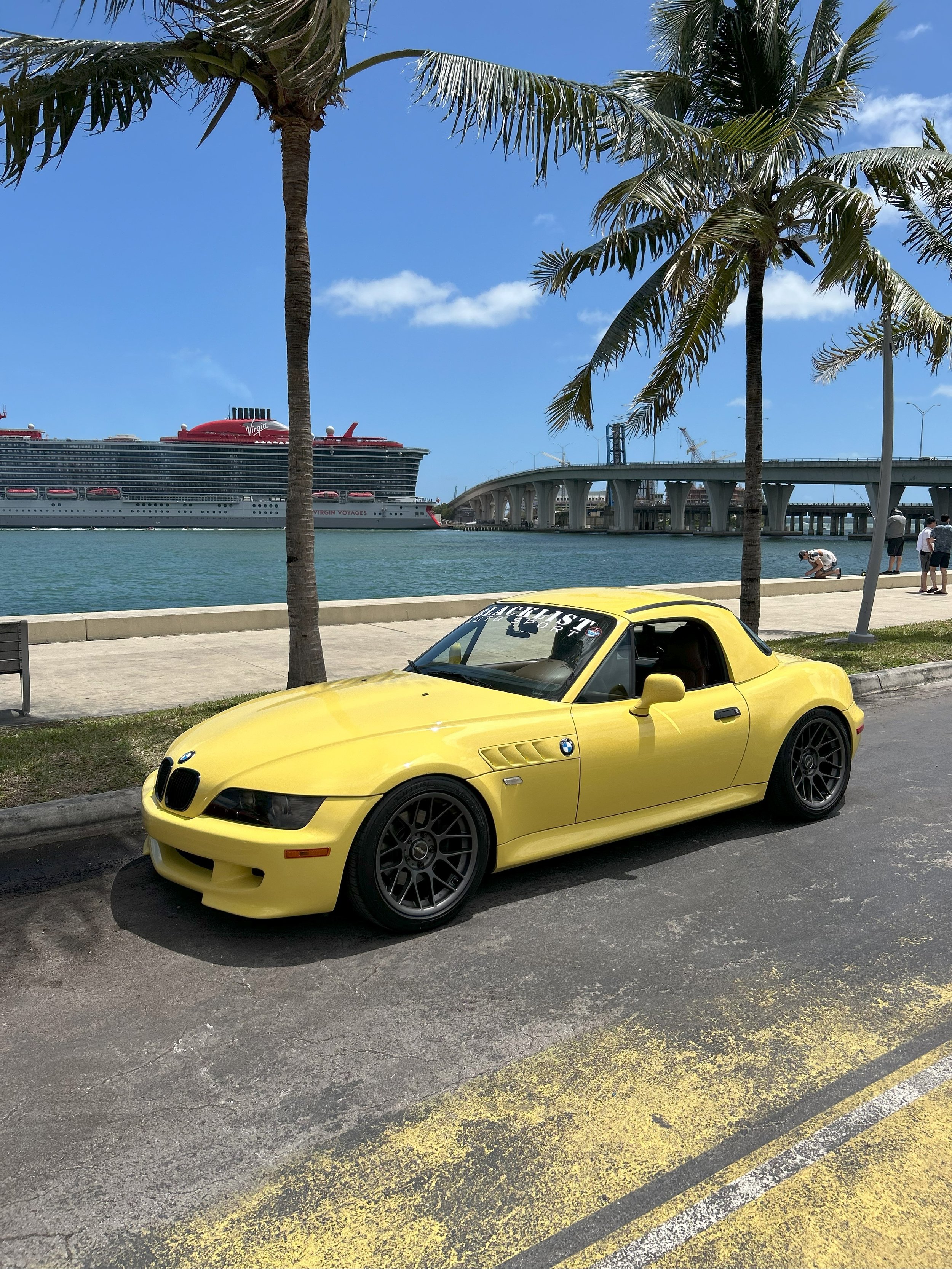 Yellow z3 bmw parked along the ocean and palm trees in miami, with a cruise ship and bridge in the background.