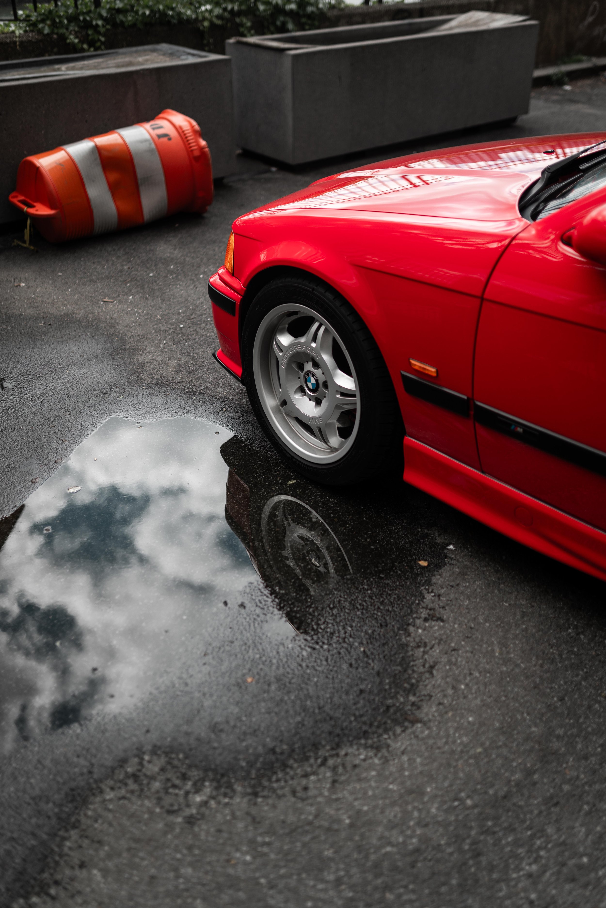 Tire detail of a 318ti tire in front of a puddle of water with the reflection of clouds in it.