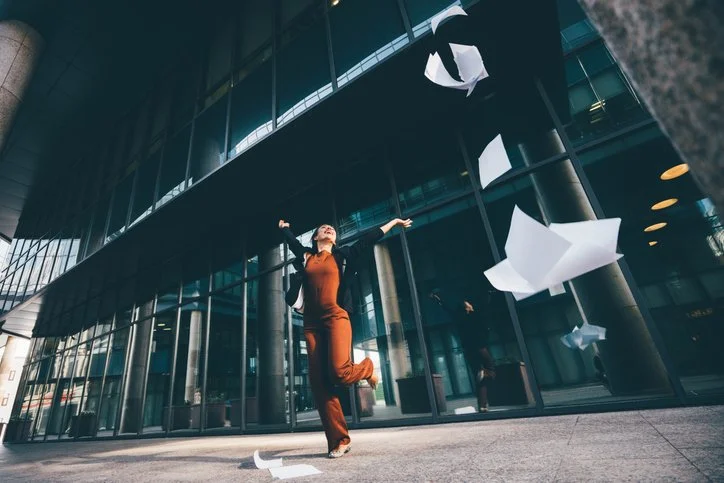 Een vrouw danst buiten een modern glazen gebouw terwijl papieren worden uitgereikt in de lucht.