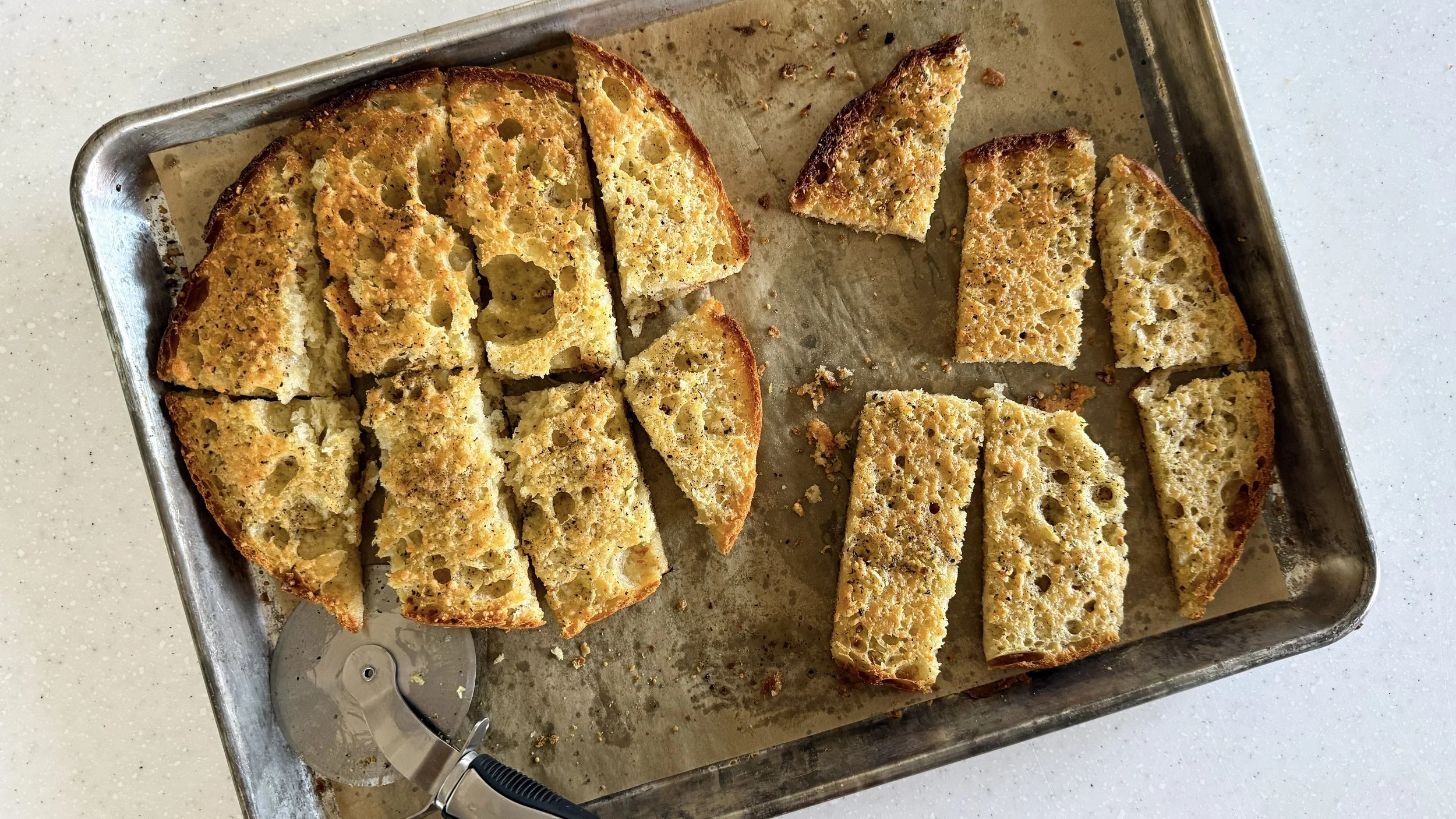 Two halves of a round focaccia loaf made into garlic bread are cut into strips of various rustic sizes on a baking sheet, with the pizza wheel resting nearby.