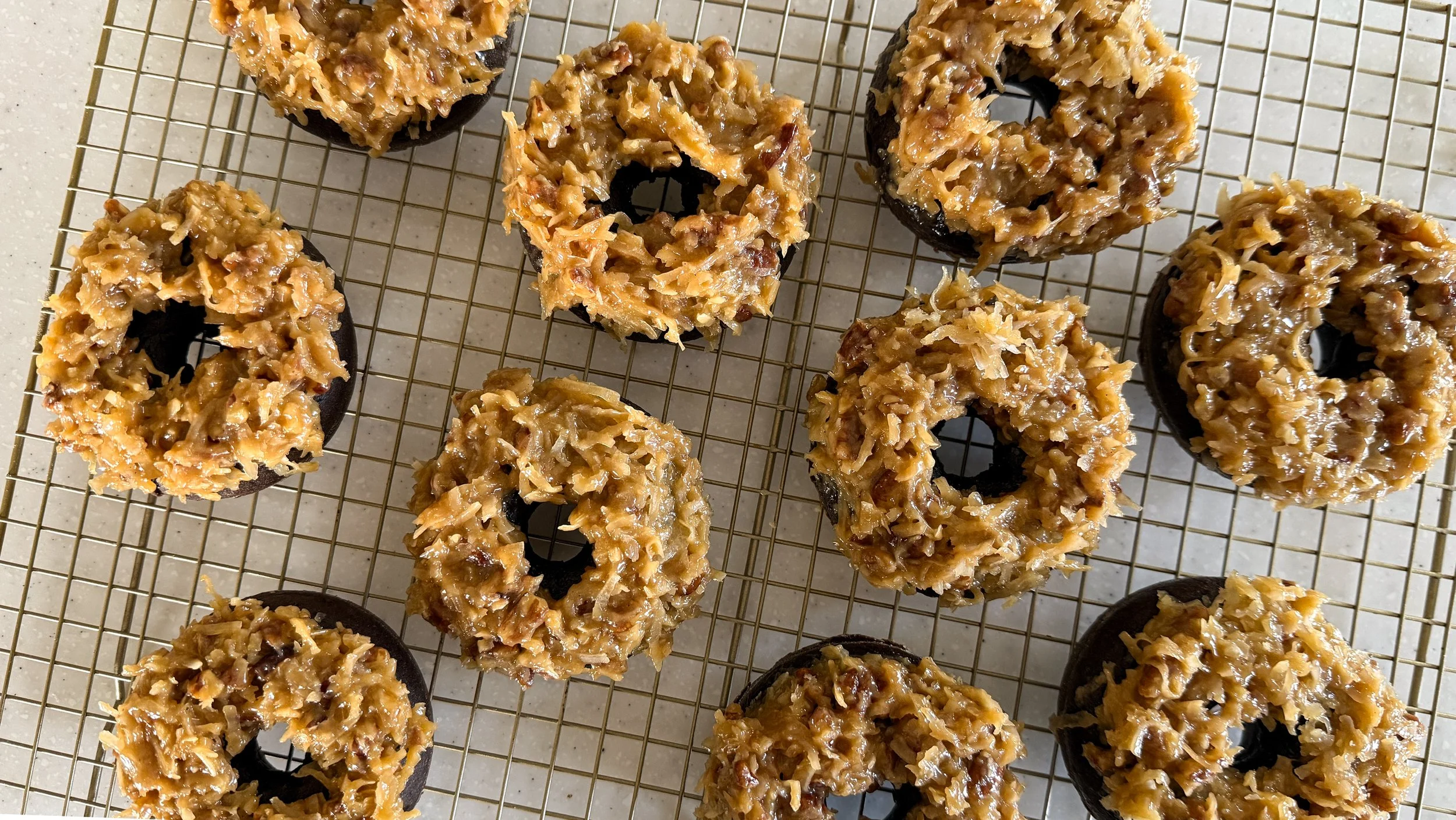 Chocolate doughnuts topped with thick coconut-pecan topping on top of a gold cooling rack.