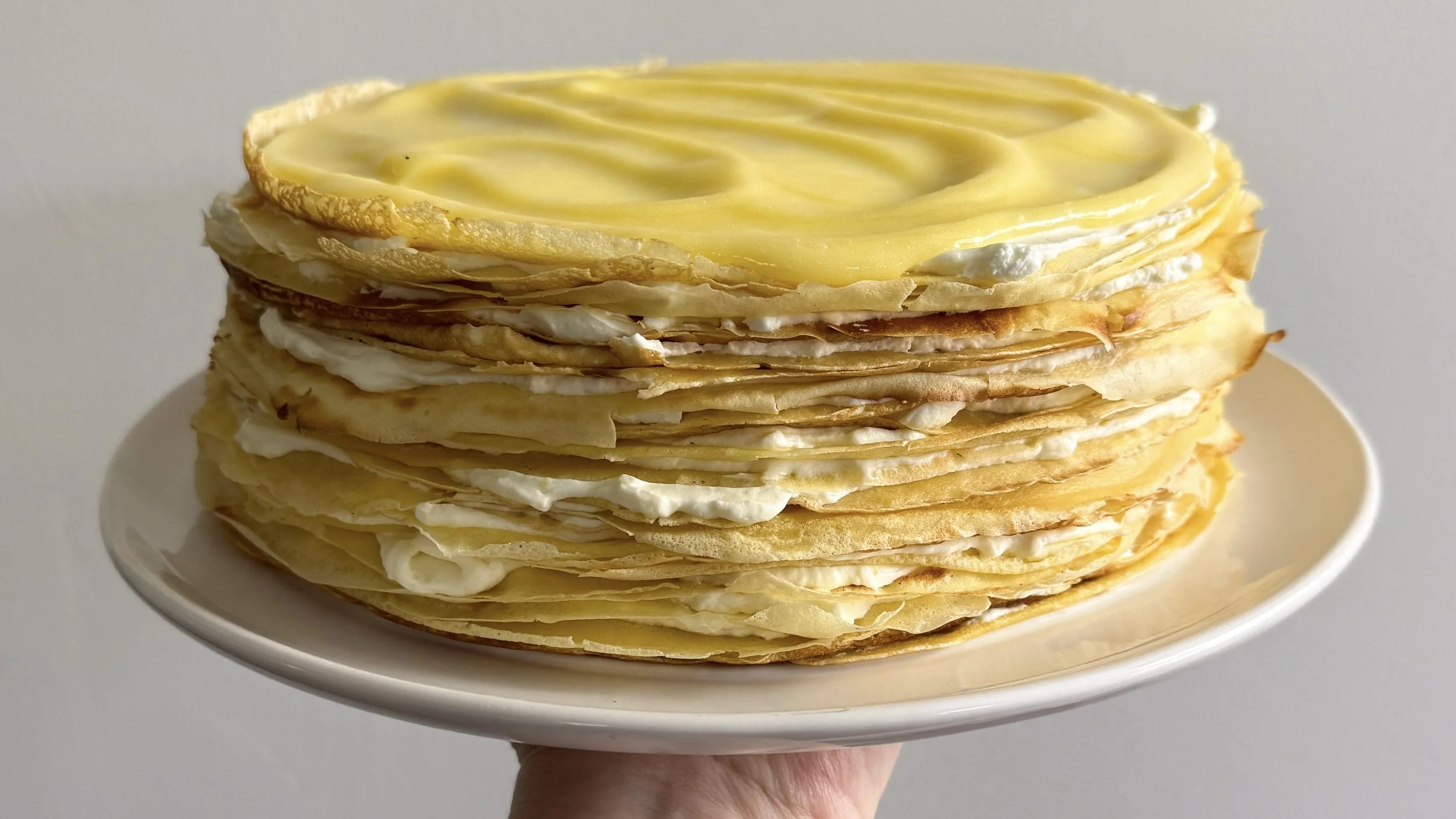 A hand holds a white plate holding a crepe cake - many layers of crepes stacked with thin layers of whipped cream, and topped with lemon curd.