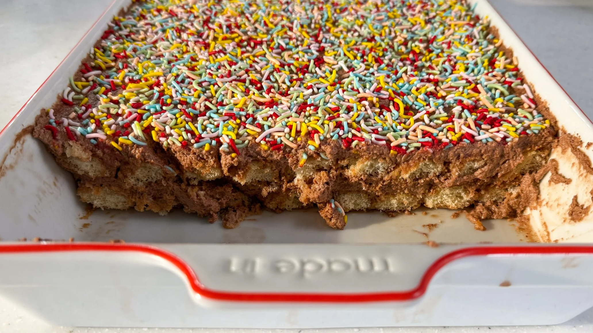 A white baking dish filled with sprinkle topped chocolate tiramisu. A portion is scooped out to show the soft and creamy interior texture.