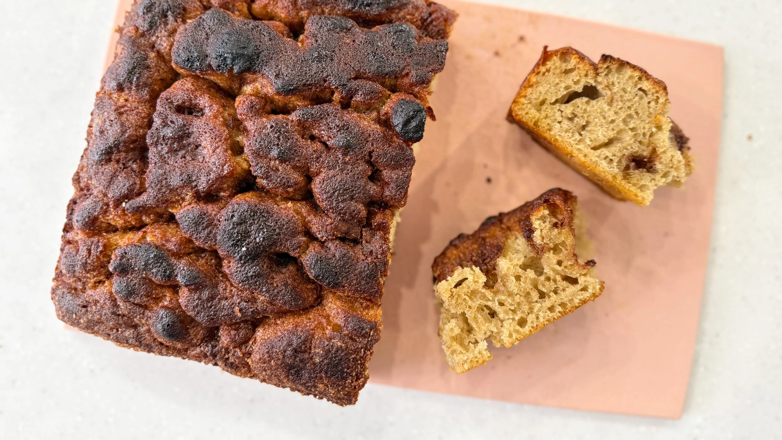 A loaf of deeply caramelized cinnamon sugar focaccia sits on a peachy colored cutting board.