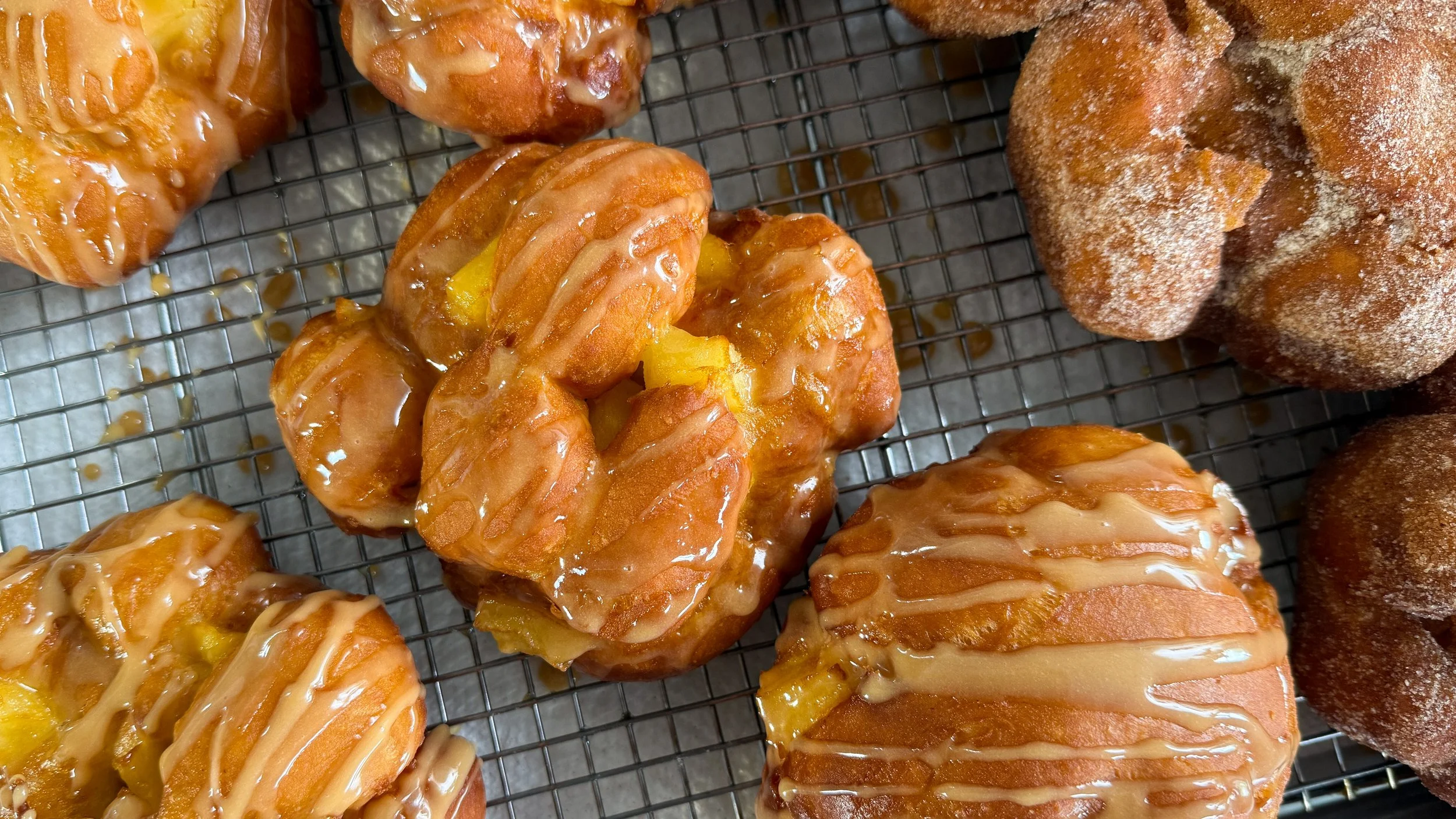 Several doughnuts on a cooling rack. Some are drizzled with glaze, and others are coated in cinnamon sugar. Visible apple pieces can be seen between pieces of the craggy dough.