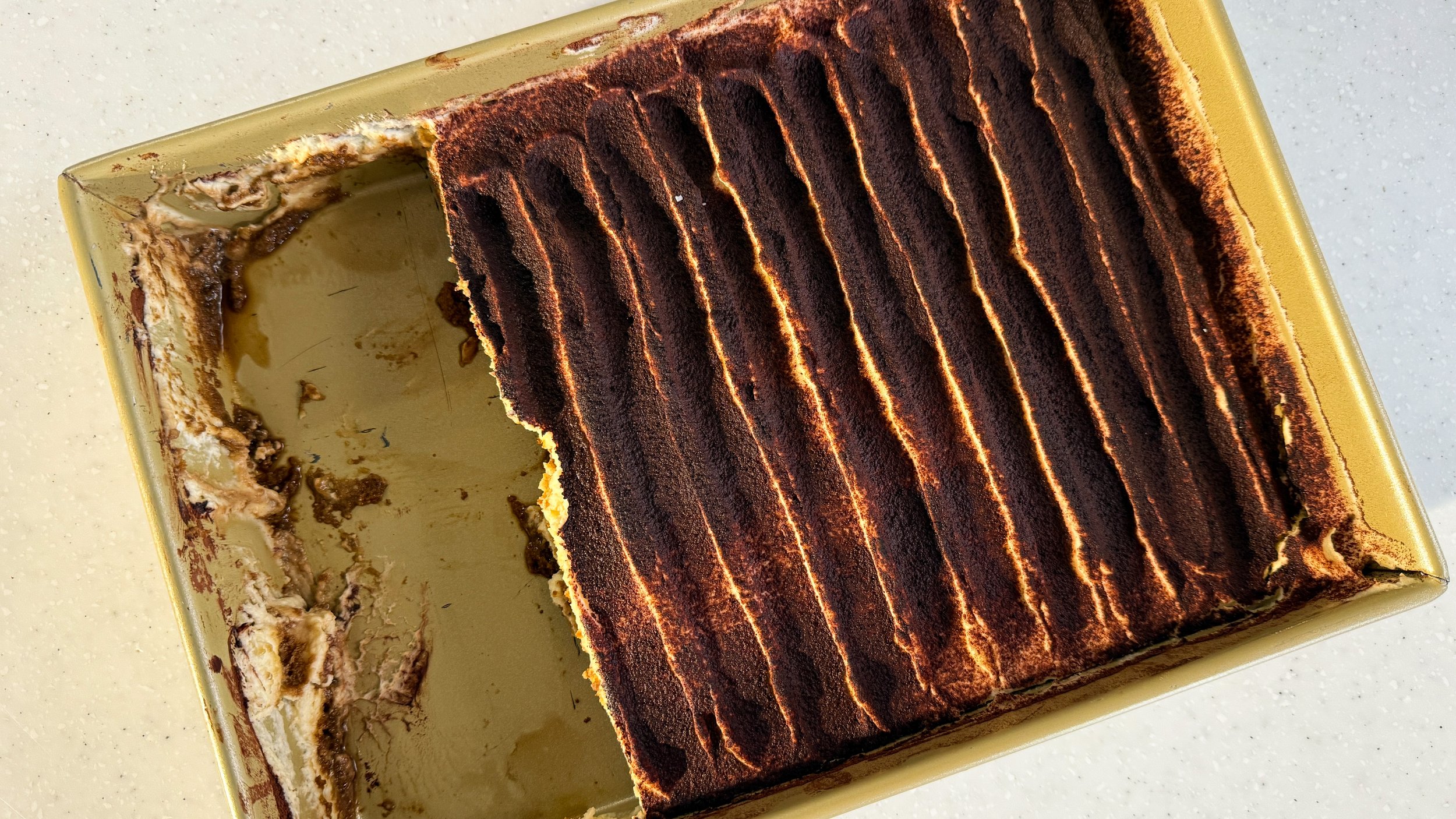 A gold baking dish filled with tiramisu - the top is textured to look sort of like stripes, and about 1/4 of the pan has been scooped out.
