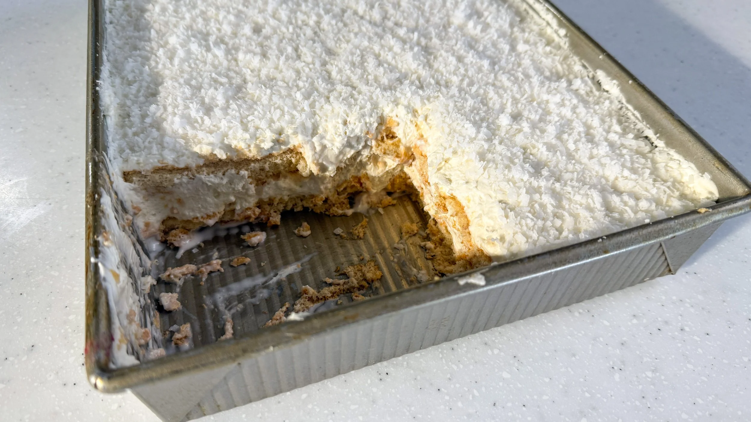A silver baking pan filled with a coconut tiramisu, part of the mixture is scooped out revealing the soft and creamy layers inside.
