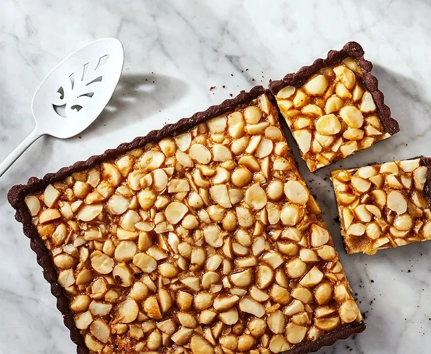 Chopped macadamia nuts and caramel in a rectangular chocolate crust with fluted edges, sitting on a marble surface with a white enamel pie server