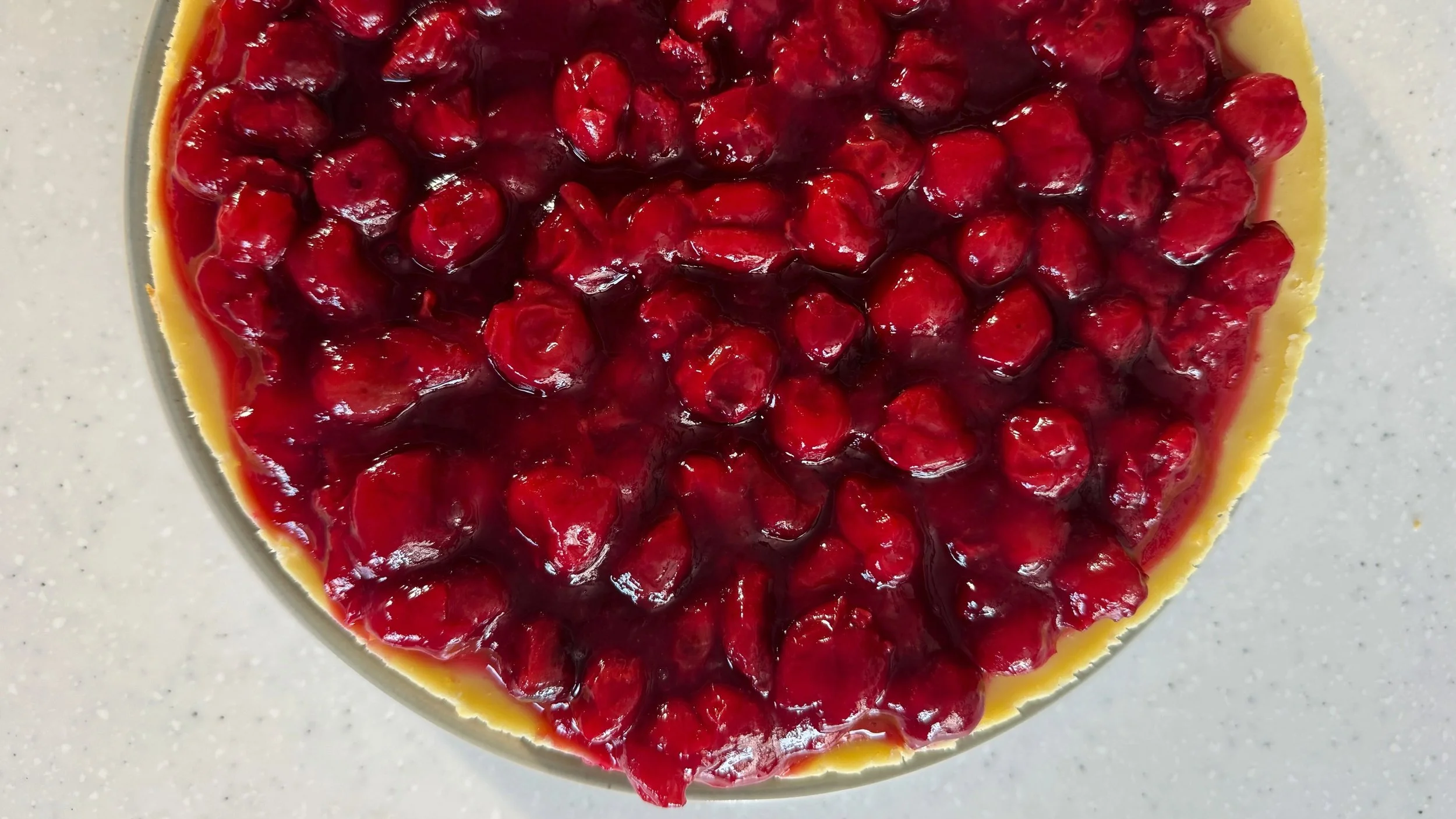 A cheesecake topped with bright sour cherry topping is shown from overhead.