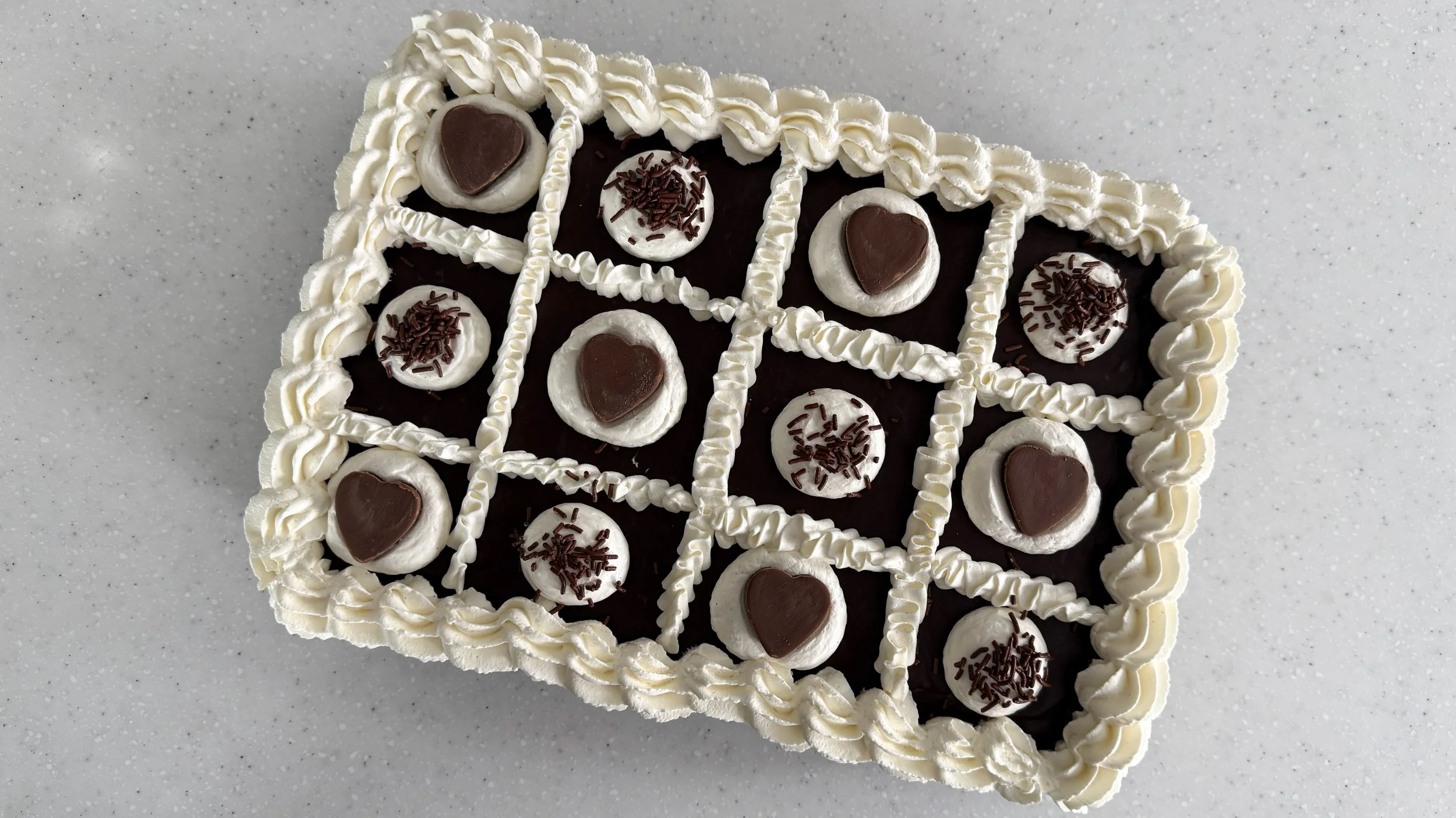 A sheet cake shown from overhead, frosted with a border of whipped cream frosting, and decorated with chocolate hearts and chocolate sprinkles.