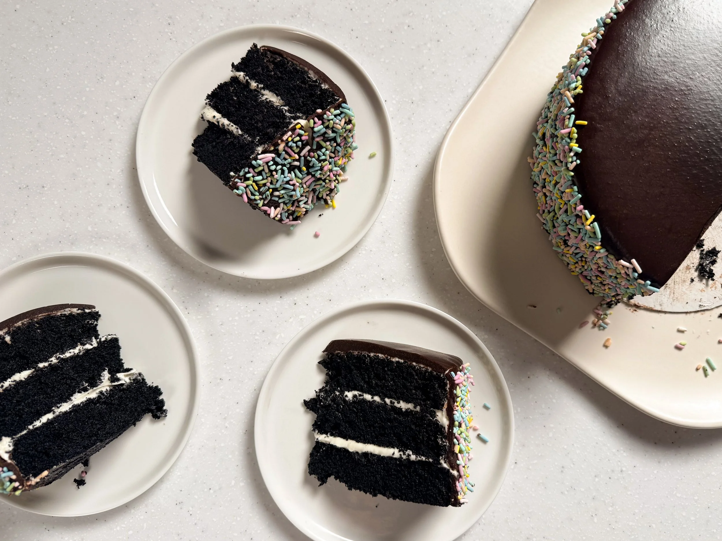 Three slices of a three layer chocolate cake filled with cream cheese frosting, glazed in fudge frosting, and covered in rainbow sprinkles. The rest of the cake peeks in on the right side.