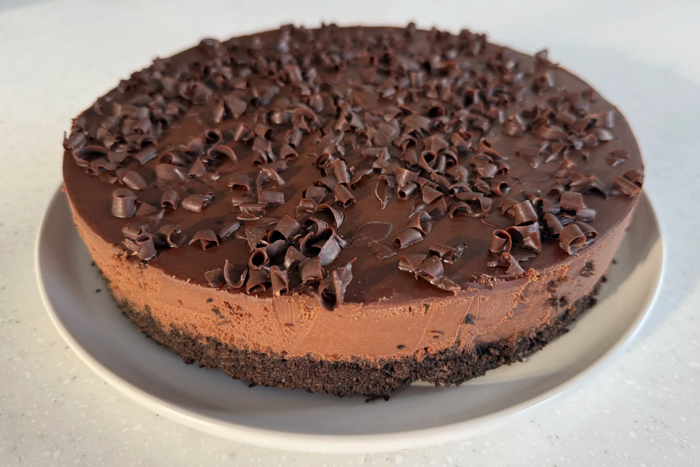 A chocolate cheesecake topped with chocolate curls on a white platter