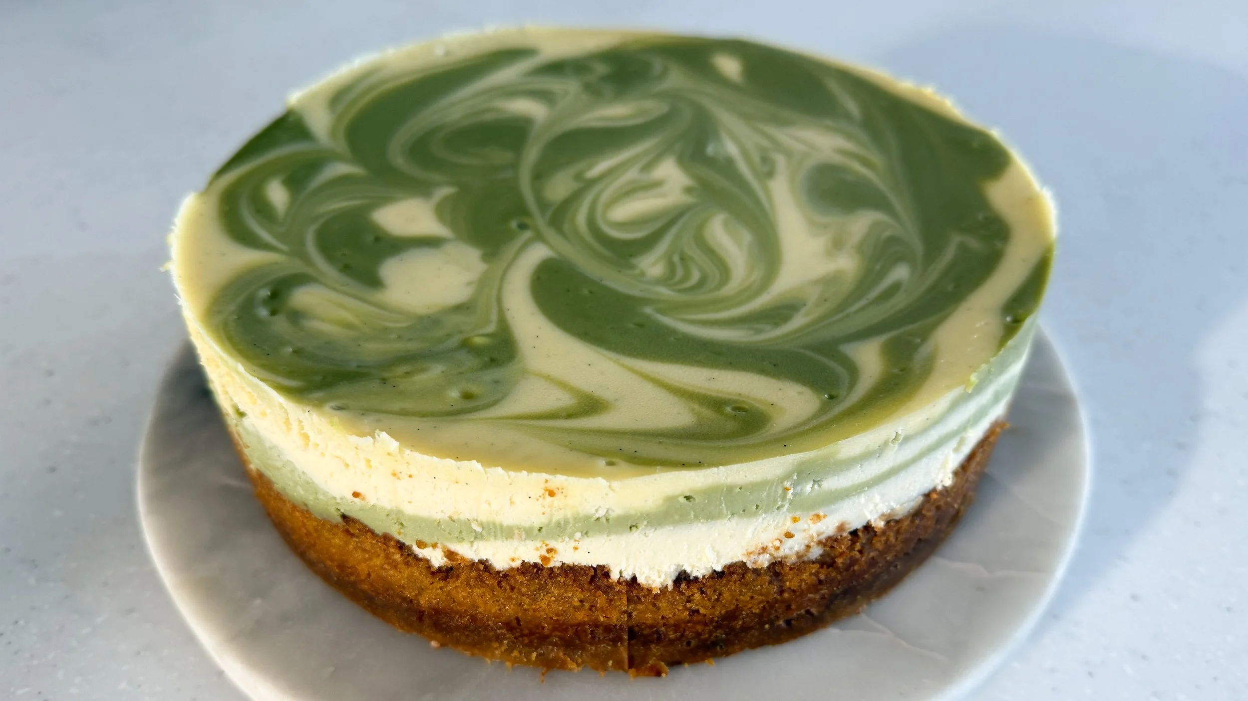 A swirly white and green vanilla pistachio cheesecake on a marble platter.