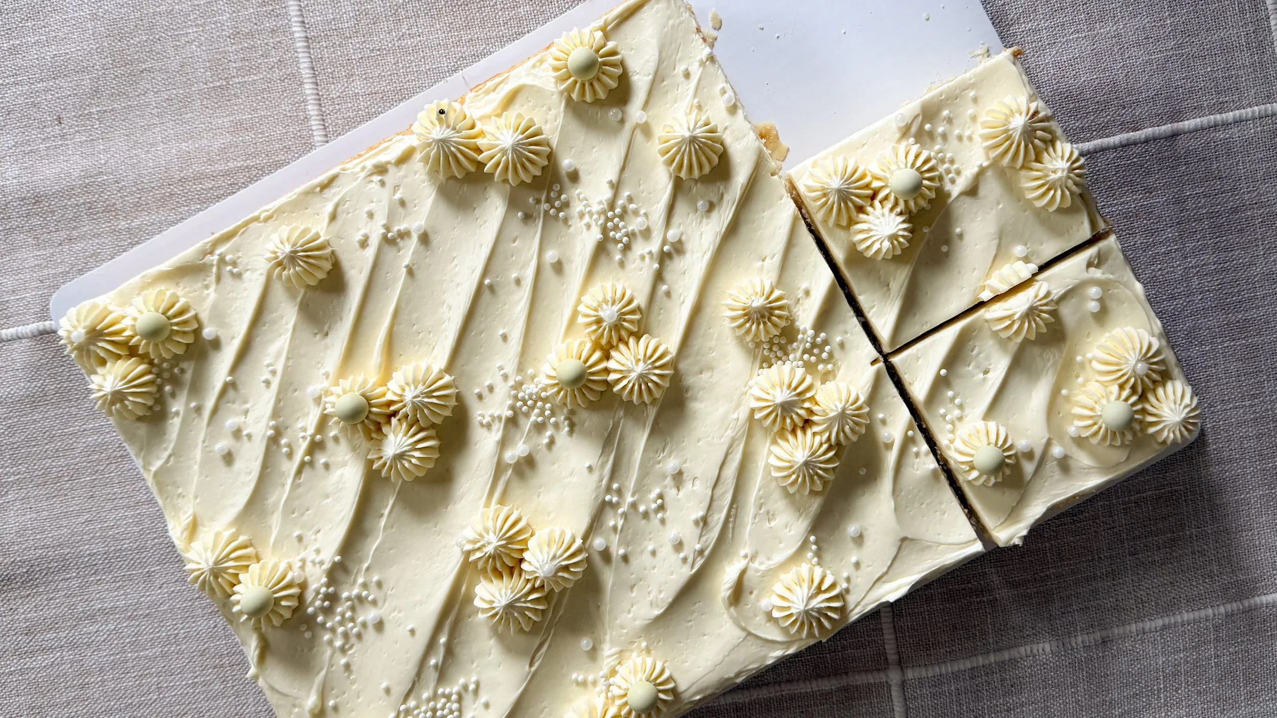 A vanilla sheet cake sits on a cloth tablecloth. It's decorated with piped stars of frosting and different sizes of white pearl and nonpariel sprinkles.