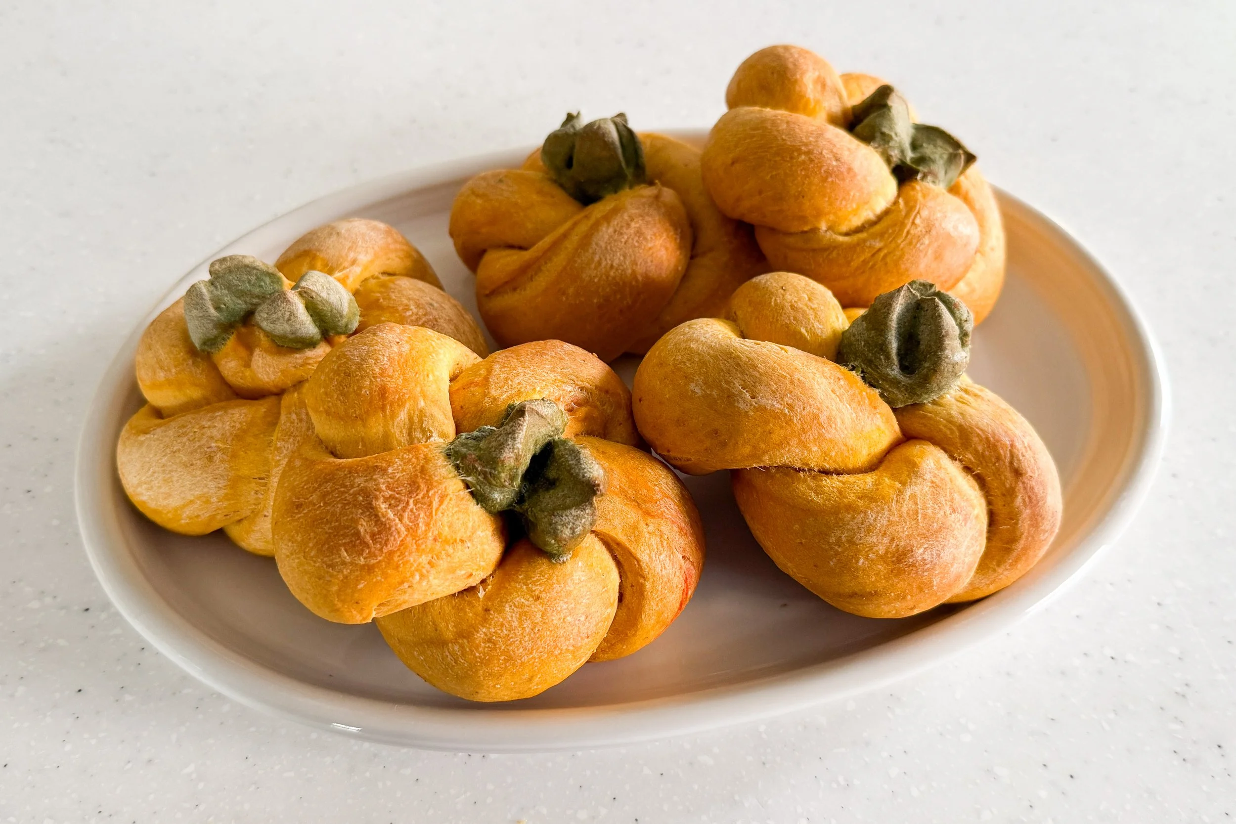 a pile of orange twisted pumpkin bagels topped with pale green stems on a white platter.