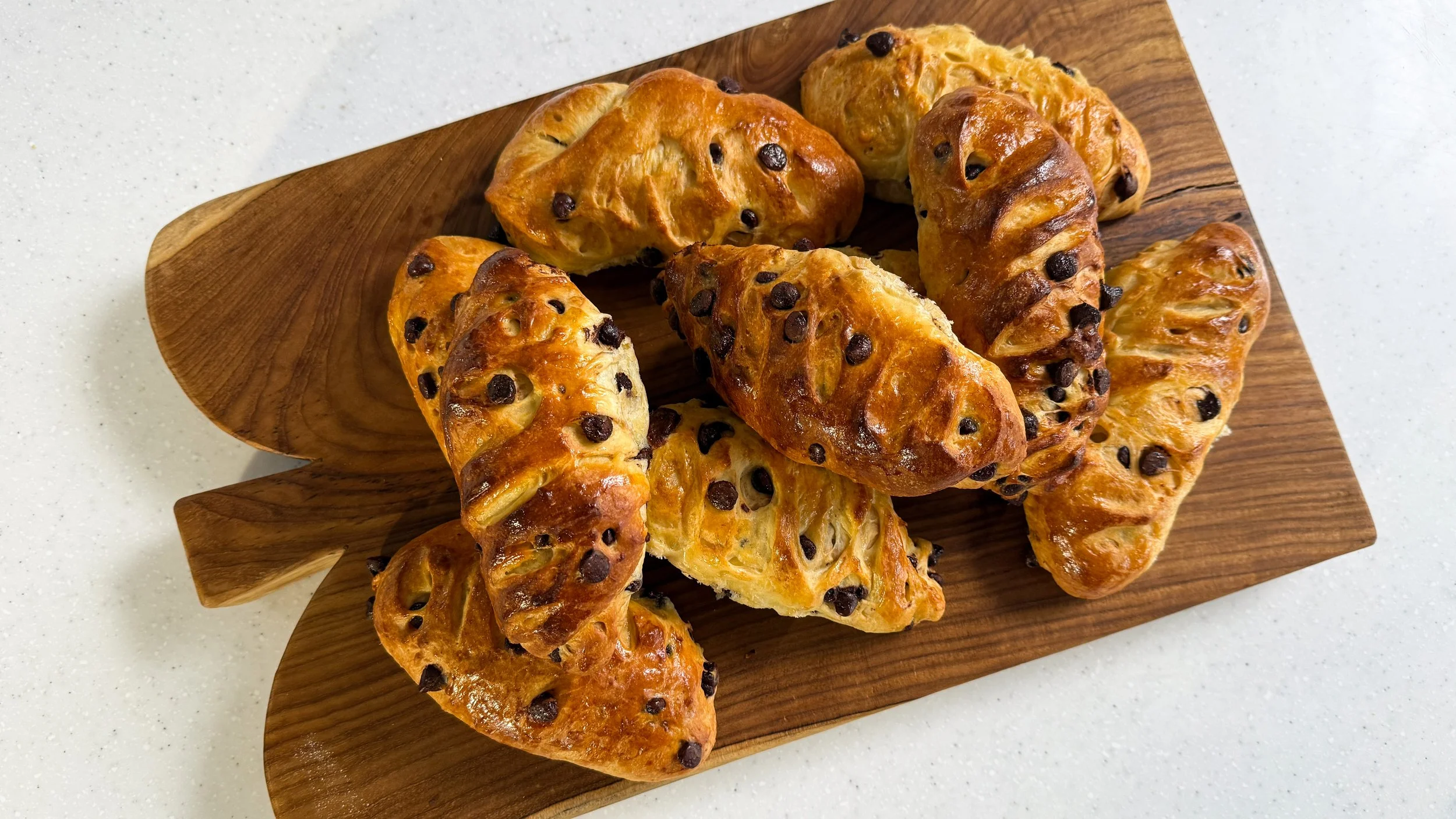 A large wooden cutting board holding a pile of golden brown, obong mini loaves studded with large pieces of chocolate.