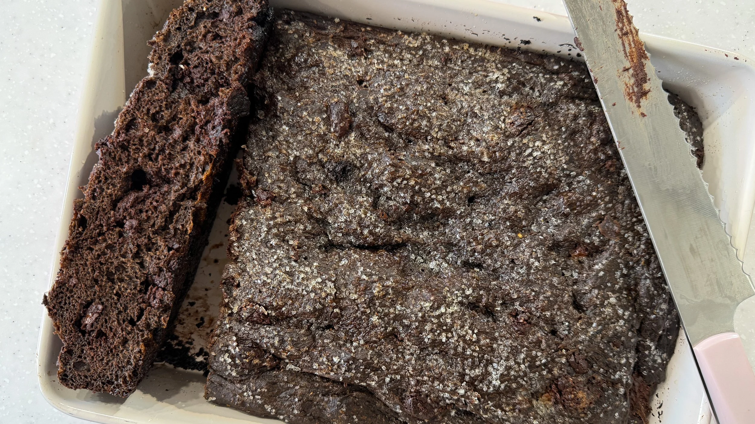 A pan of chocolate chunk focaccia topped with sandwing sugar is in a pan, one long slice has been removed and turned upward to show the fluffy interior crumb.