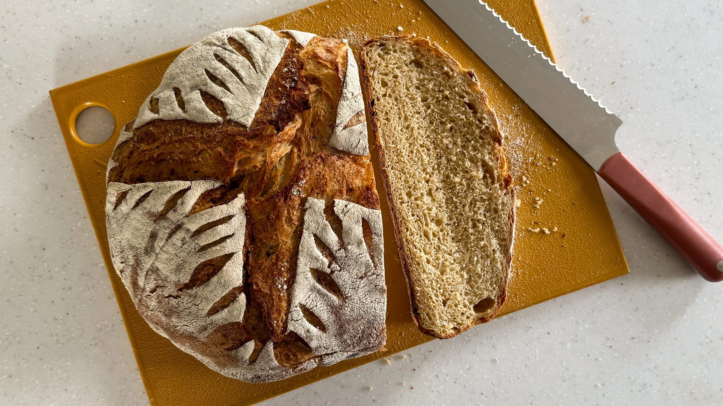 A loaf of bread with a golden crust and an intricately scored pattern on the floured surface sits on a golden cutting board, with a pink handled serrated knife to the right.
