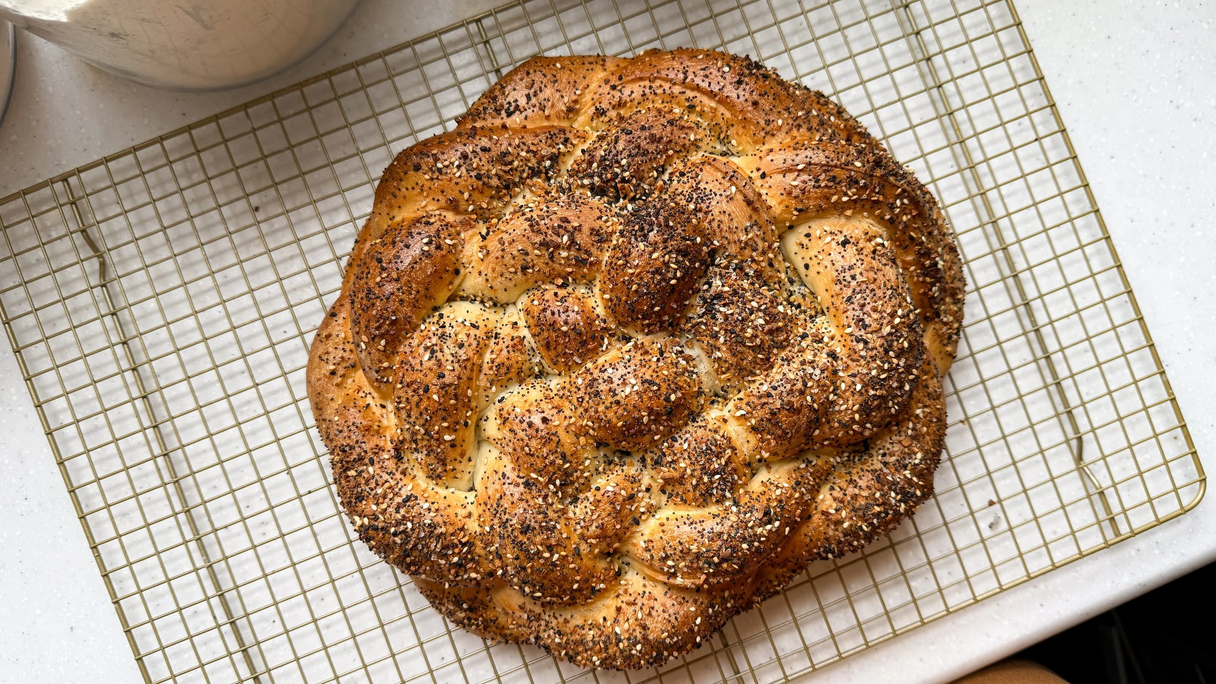 A braided loaf of pretzel bread topped with everything bagel seasoning sits on a cooling rack.