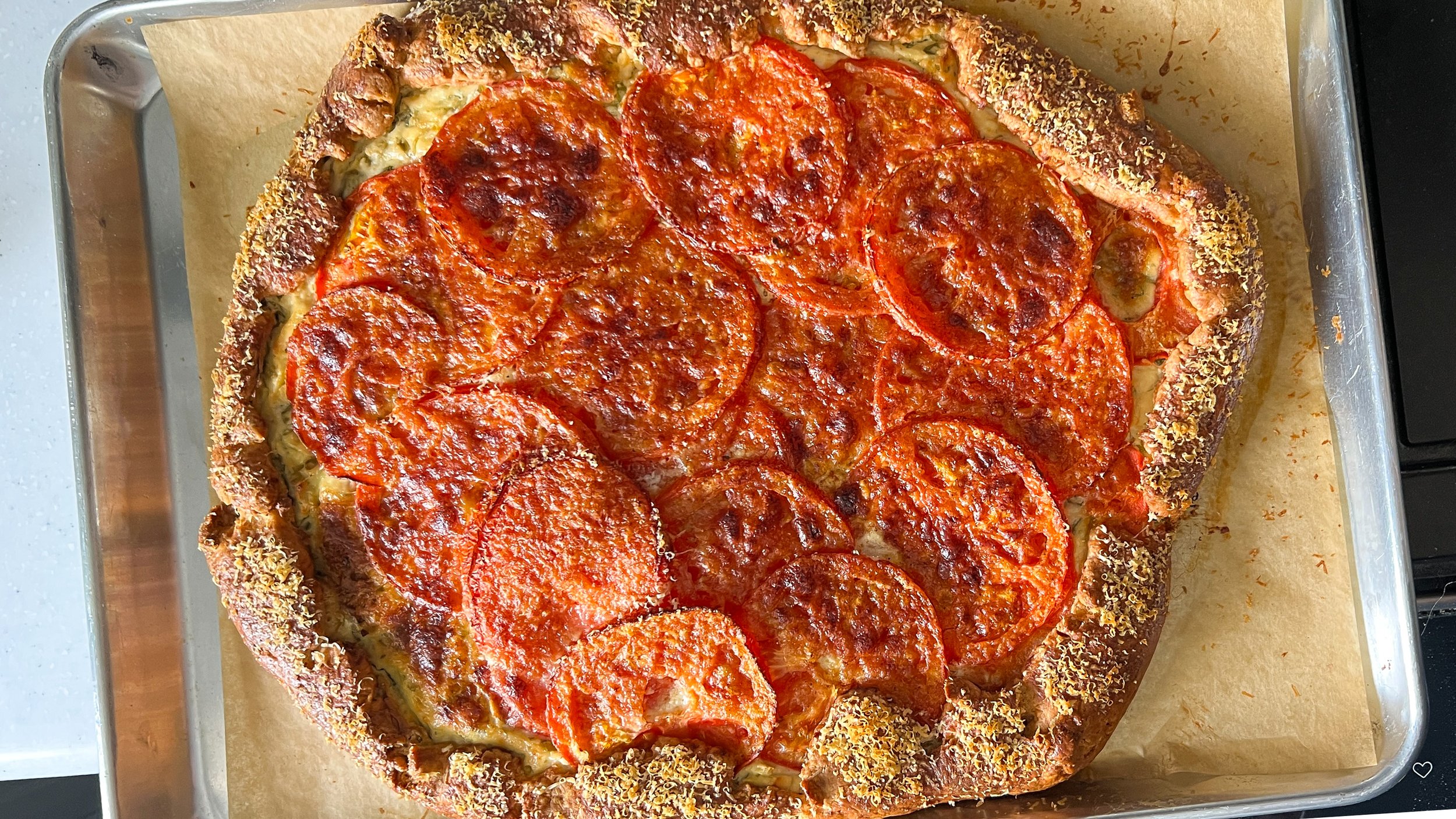 A parmesan-crusted edge encases a galette fitted with sliced tomatoes on a parchment lined baking sheet.