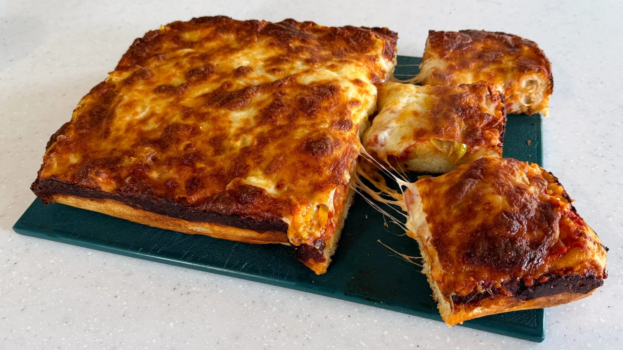 A rectangular loaf of focaccia topped like pizza sits on a green cutting board. Three pieces have been cut, and two have been partially pulled away, showing strings of melty cheese.