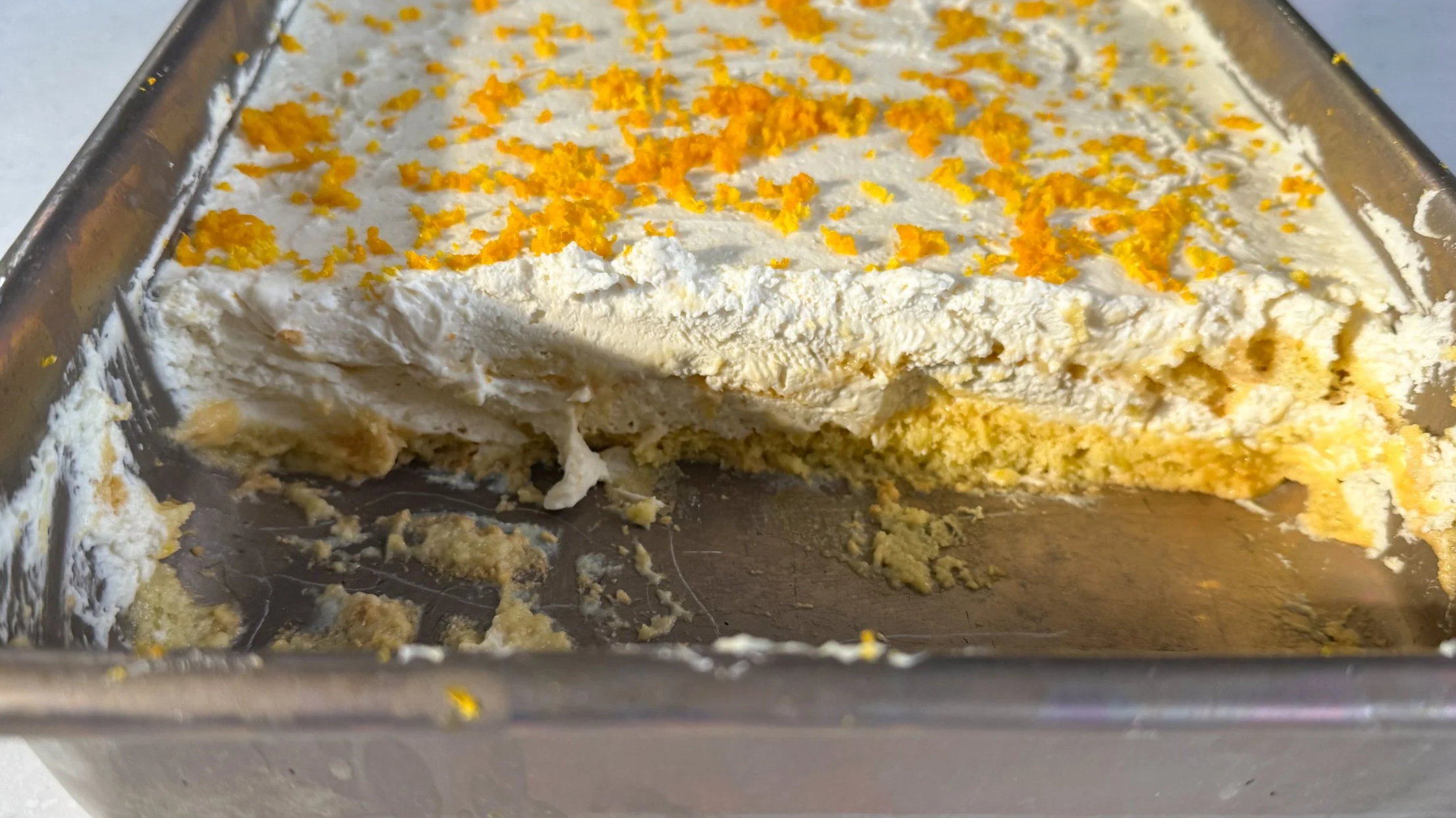 A silver baking pan filled with an orange-tinted tiramisu, topped generously with orange zest. About 1/4 of the pan is scooped out to reveal the creamy, soft interior texture.
