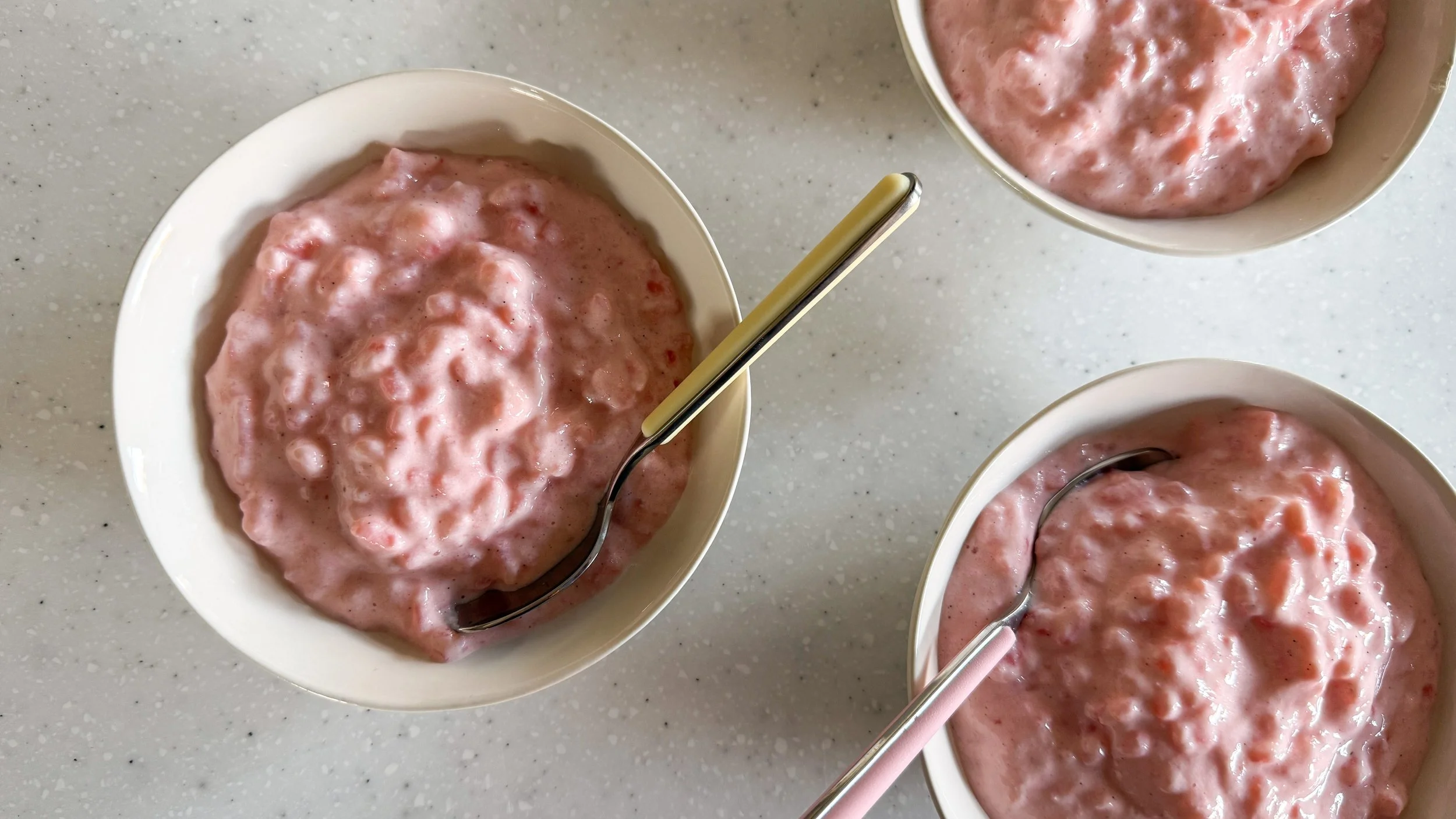 Three white bowls of pink rice pudding sit on a white kitchen counter. One bowl has a yellow spoon resting in it, and another has a pink spoon.