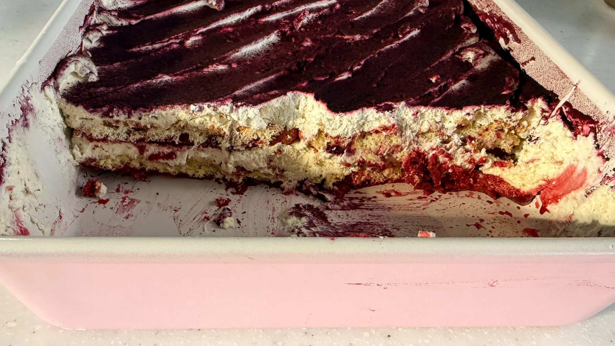 A pink baking dish filled with a pink layered tiramisu made with hibiscus tea. About 1/4 of it is scooped out to reveal the soft and creamy internal texture.