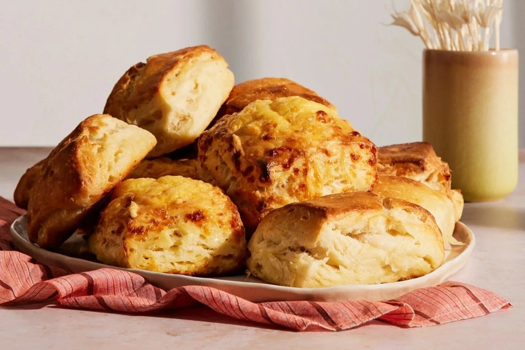 A pile of tall, golden brown biscuits on a white plate sitting on top of a rust colored striped napkin. A vase of dried flowers is visible in the background.