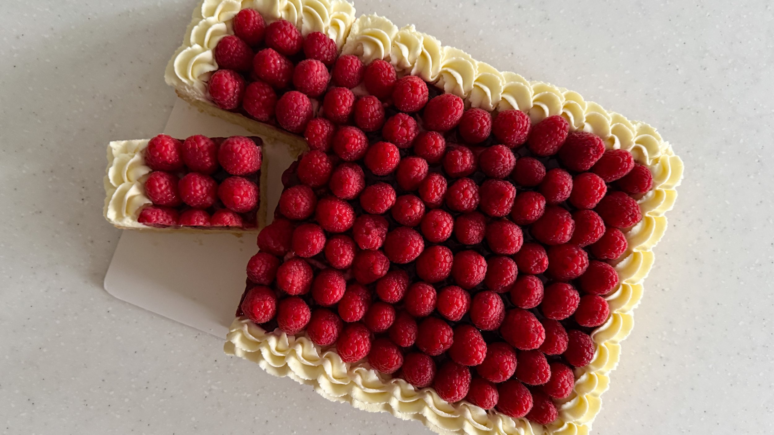 A sheet cake shown from overhead, frosted with a border of lemon frosting, a layer of jam, and fresh raspberries fully covering the surface.