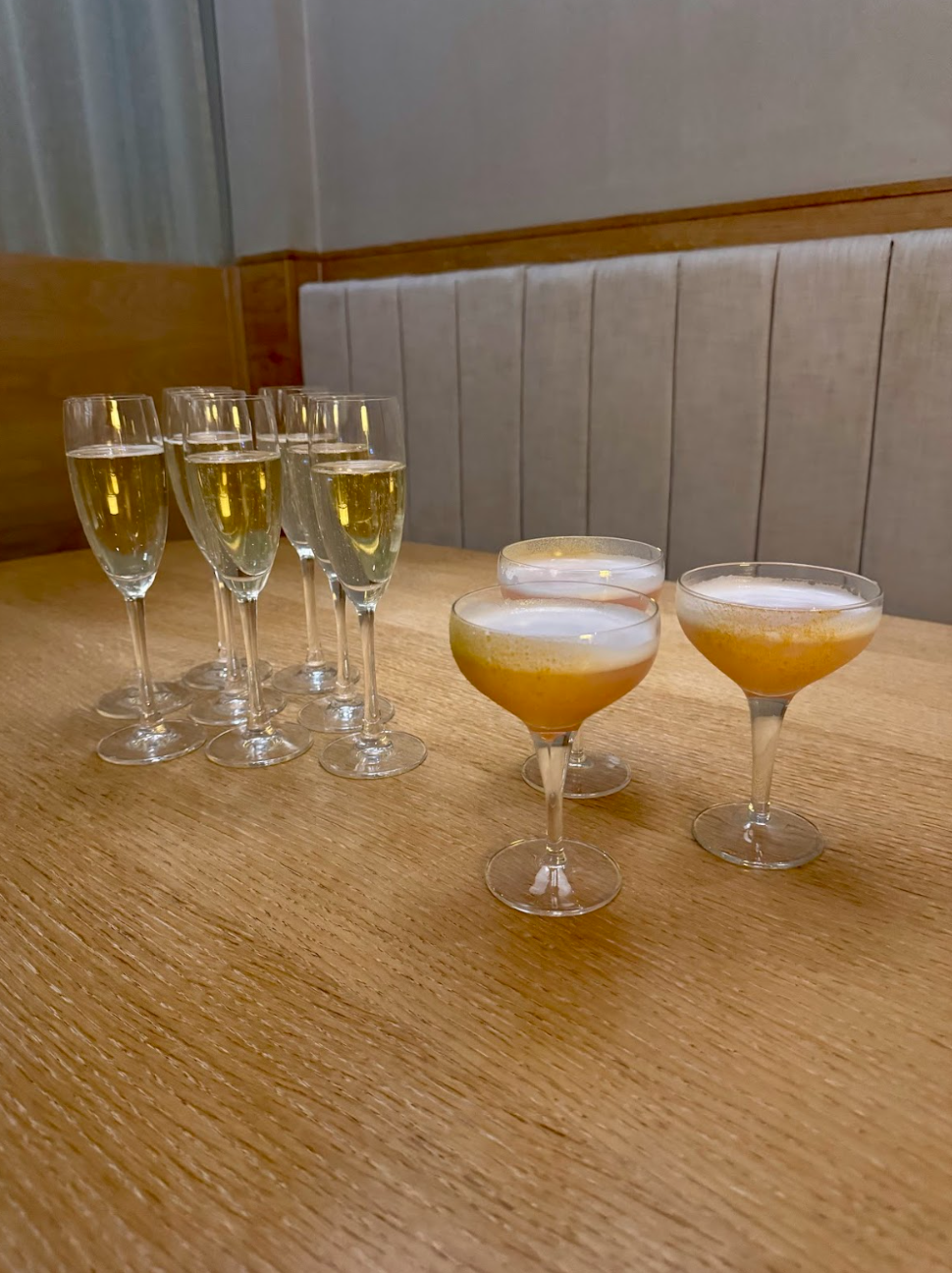 Six champagne flutes filled with sparkling wine and three cocktail glasses with yellow and white layered drinks on a wooden table.