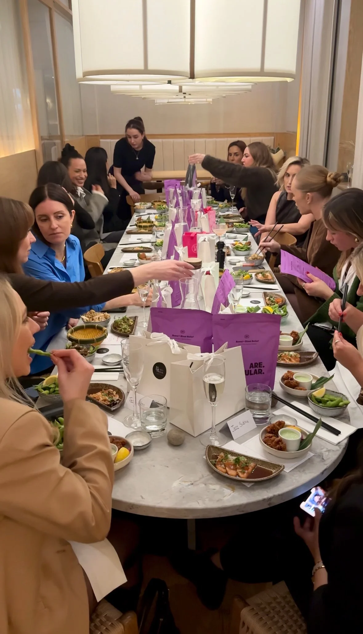 Group of women sitting at a long dinner table during a gathering or celebration, with food, drinks, and gift bags.