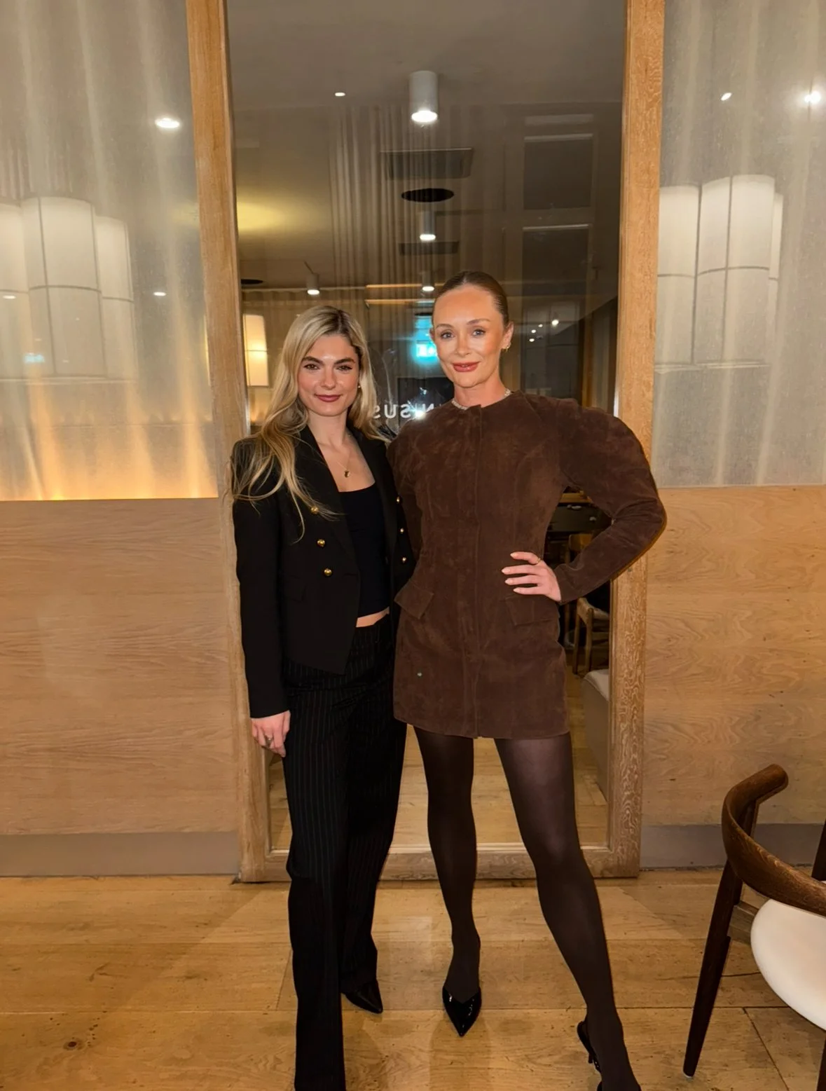 Two women standing inside a restaurant near a wooden wall, one wearing a black blazer and pants, the other in a brown dress with black tights and heels.