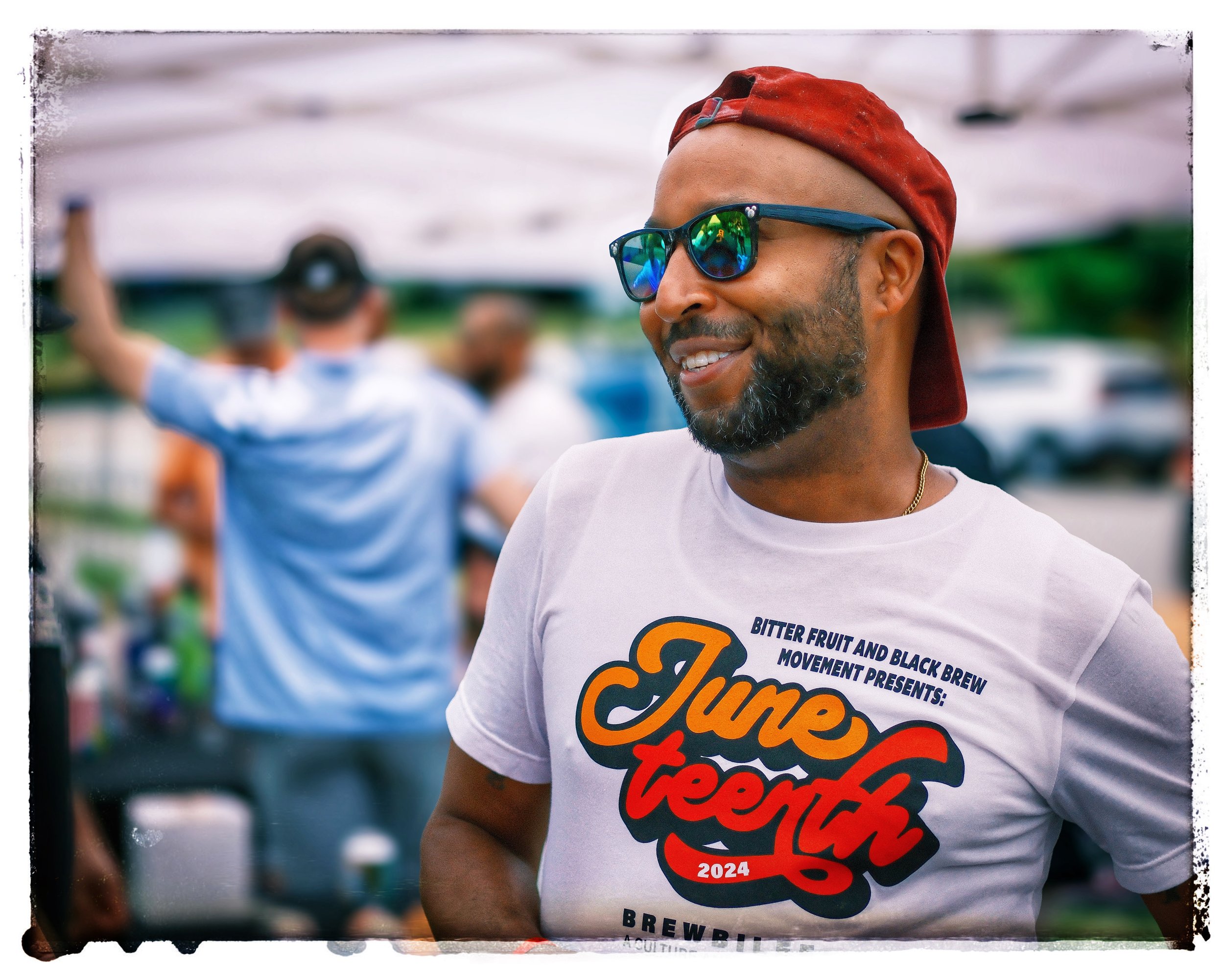 Man wearing sunglasses and a red cap backward, smiling and wearing a "Juneteenth 2024" T-shirt at an outdoor event, with people in the background.