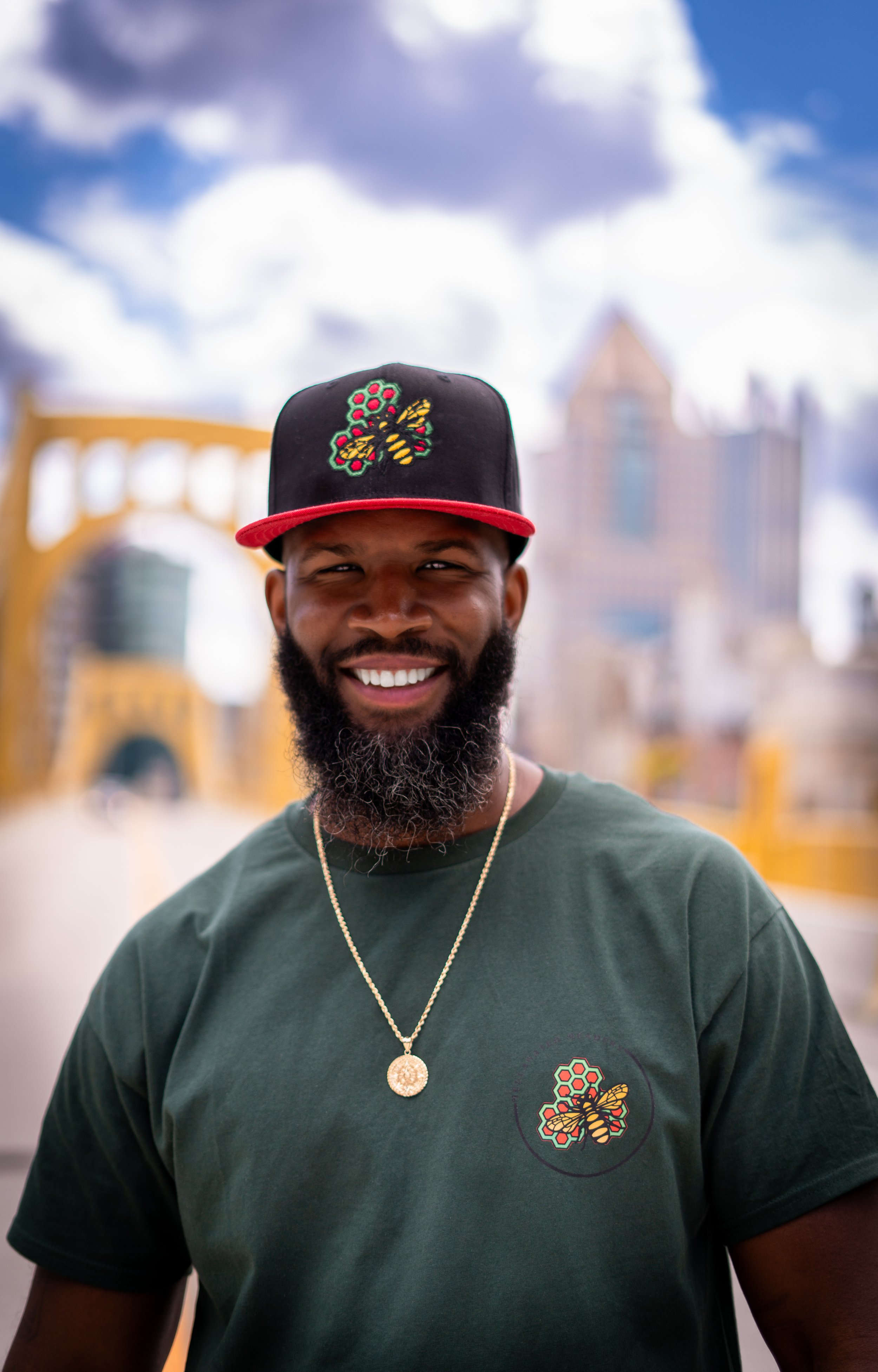 Man smiling, wearing a dark green shirt and a black and red cap with a bee design in an urban setting with a bridge in the background.