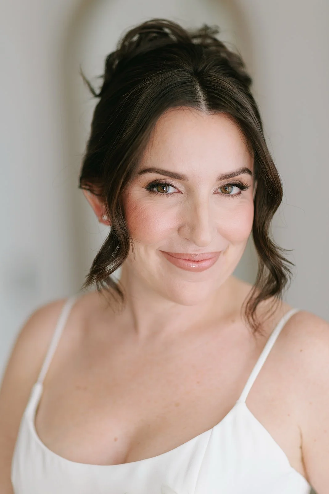 Close-up of a smiling woman with brunette hair styled in loose waves, wearing a white spaghetti strap top, and looking slightly to the side.