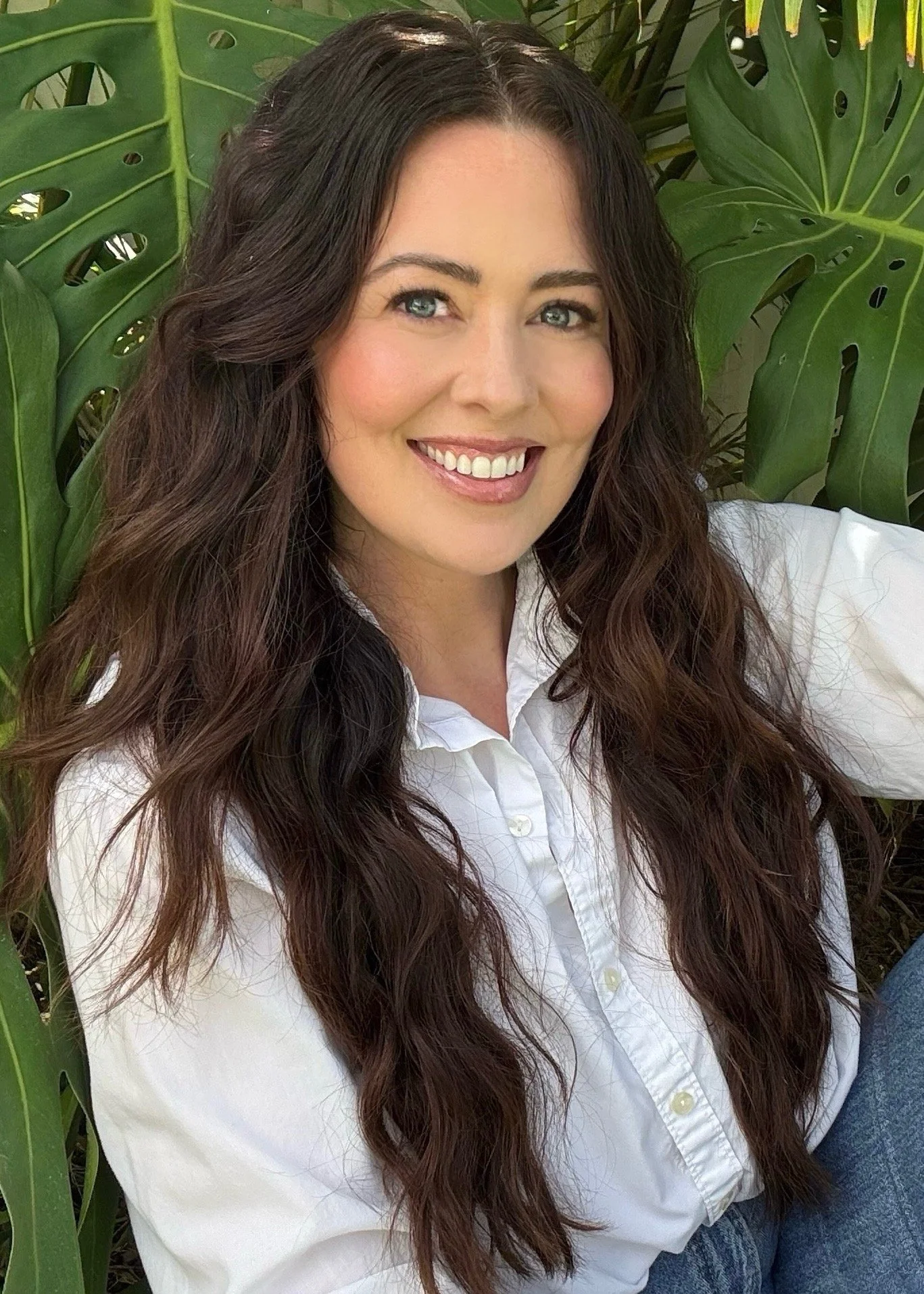 A woman with long wavy brown hair, blue eyes, and fair skin, smiling, wearing a white button-up shirt, sitting among green leafy plants.
