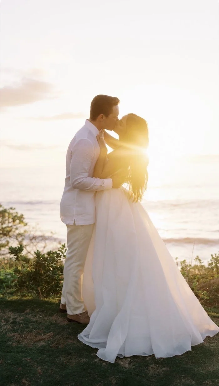 A couple kissing on a beach at sunset, with the sunlight shining behind them.