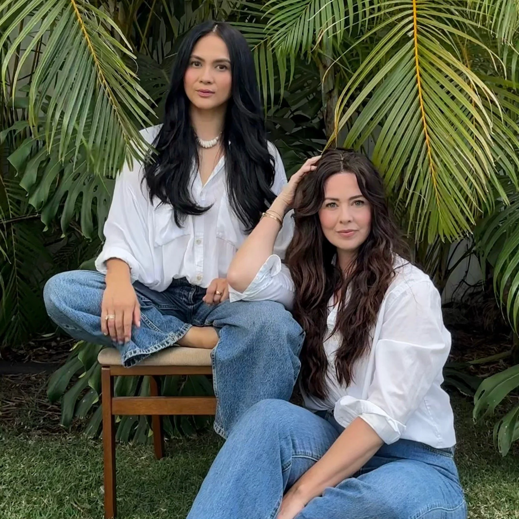 Two women with long wavy hair, one with black hair sitting cross-legged on a chair and the other with brown hair sitting on the grass, both wearing white shirts and blue jeans, posing outdoors with lush green tropical plants in the background.